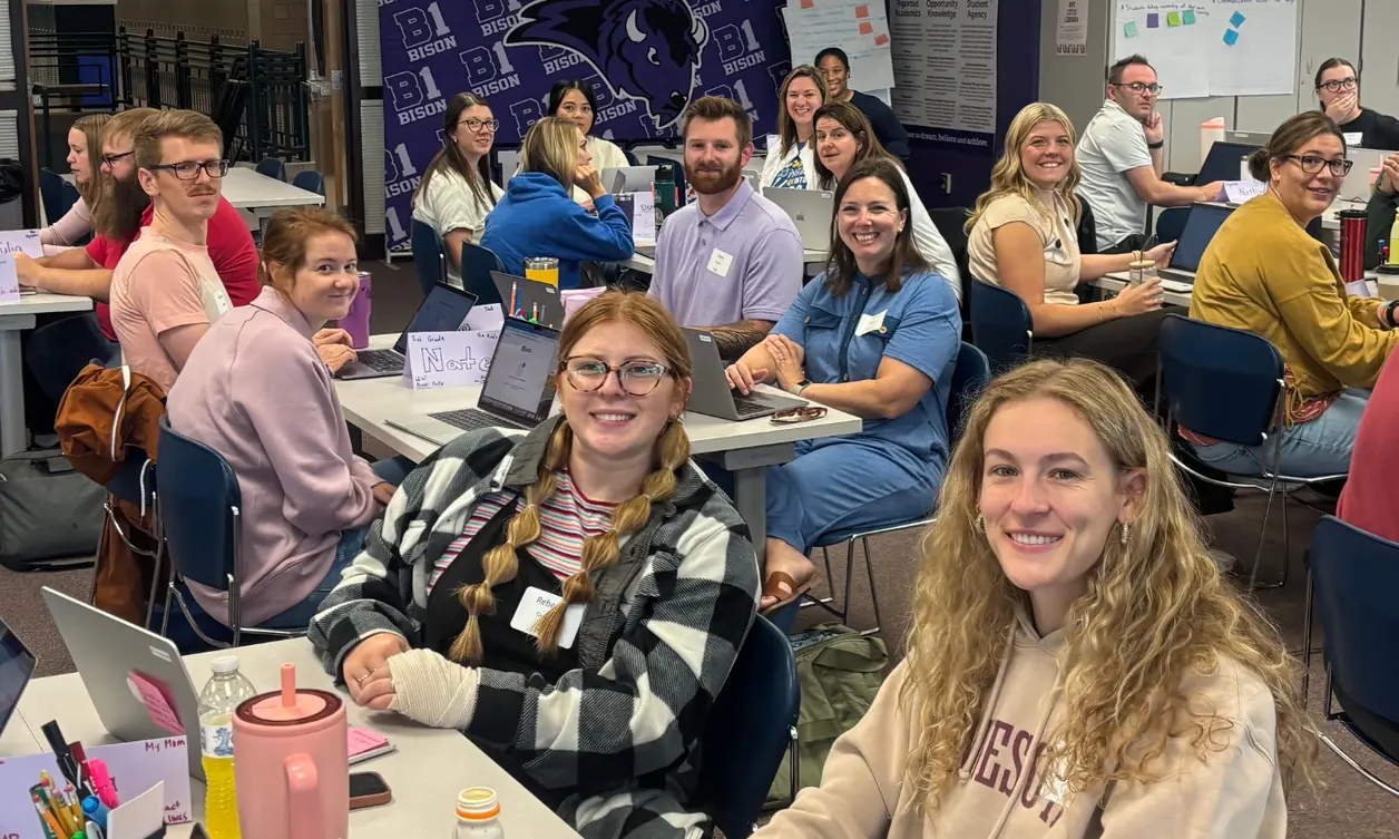 Group of people sitting at tables with laptops and notebooks, smiling and looking at the camera in a classroom or meeting room setting. Group of people sitting at tables with laptops and notebooks, smiling and looking at the camera in a classroom or meeting room setting.