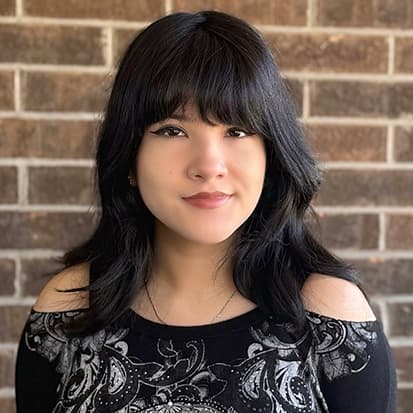 Image of a person with long black hair wearing a black and white patterned top, photographed against a brick wall background. Image of a person with long black hair wearing a black and white patterned top, photographed against a brick wall background.