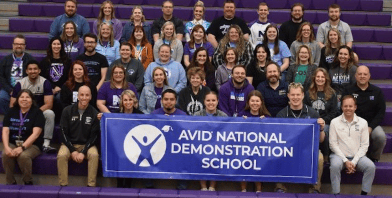 Group photo of people sitting on bleachers holding a banner that reads 'AVID National Demonstration School'. Group photo of people sitting on bleachers holding a banner that reads 'AVID National Demonstration School'.