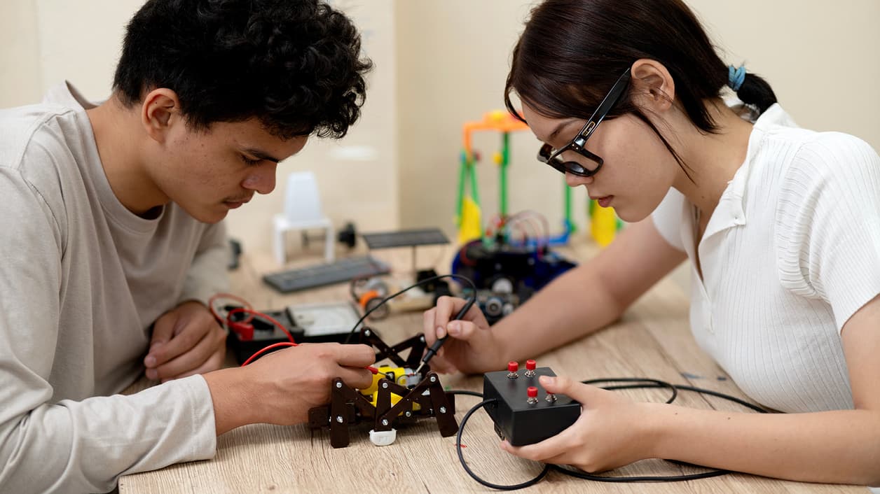 Two individuals working on a robotic project with various electronic components on a wooden table. Two individuals working on a robotic project with various electronic components on a wooden table.