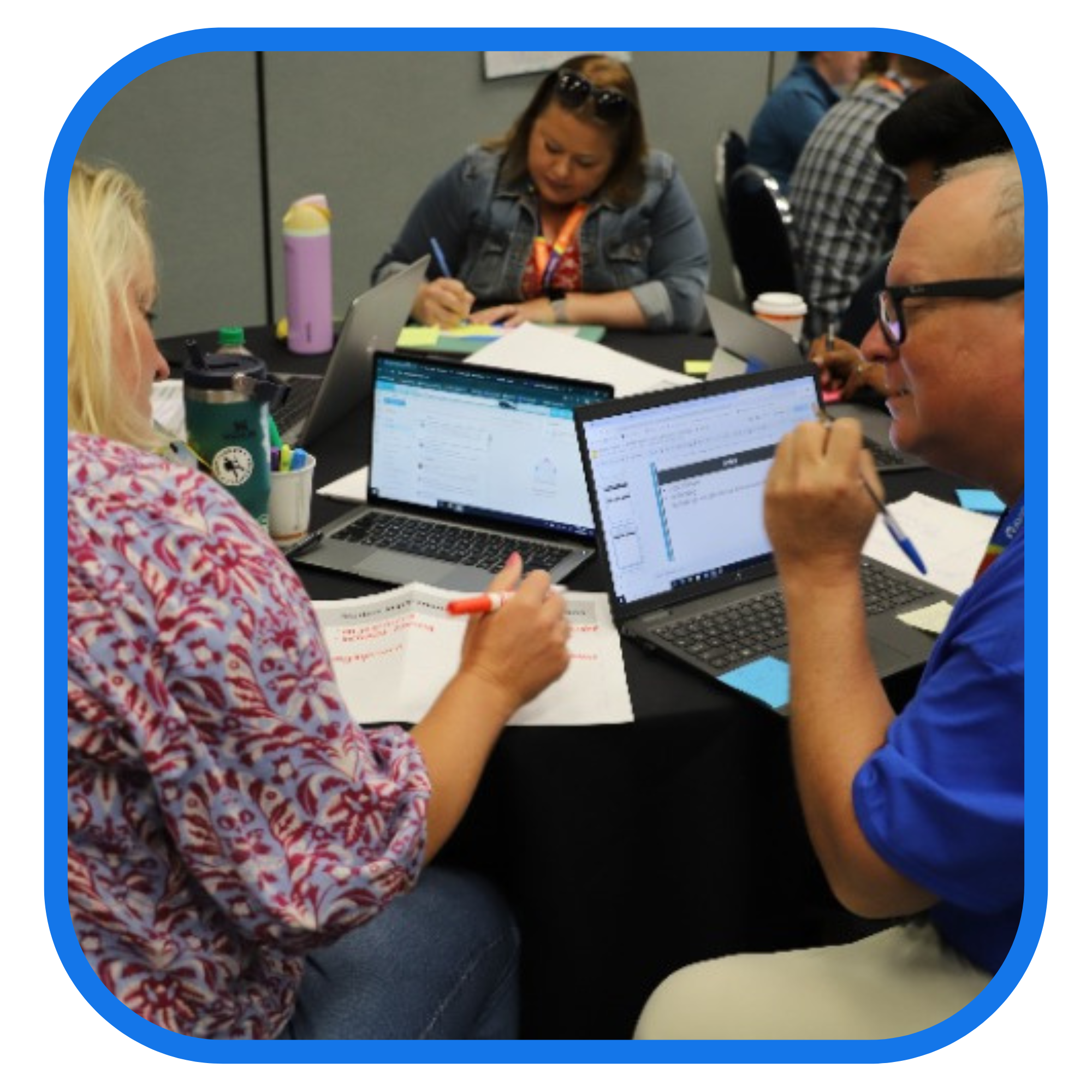 A group of people in a meeting or collaborative work setting, each with a laptop, writing on papers and discussing.