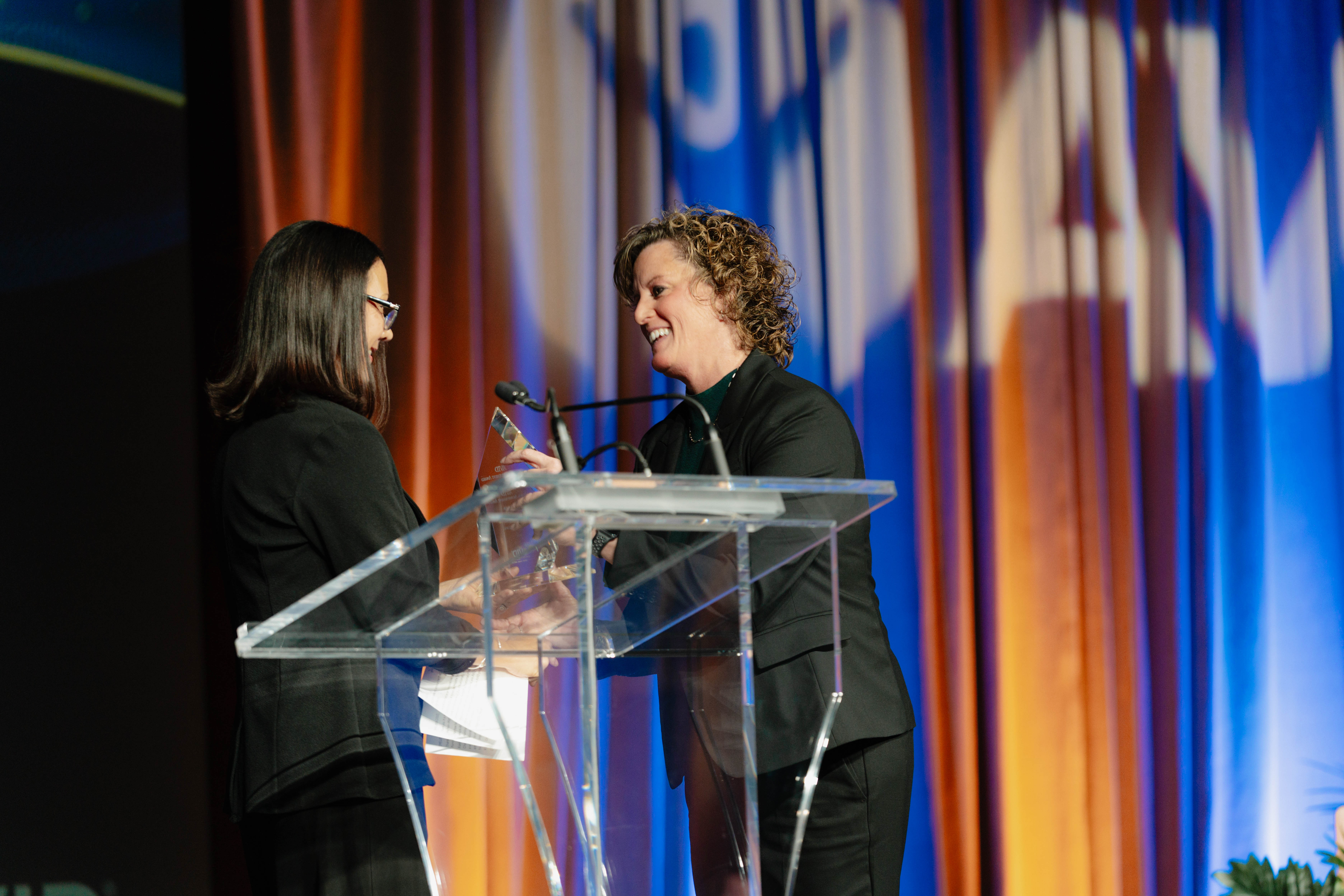Two individuals standing at a podium on a stage with colorful curtains in the background, engaging in conversation.