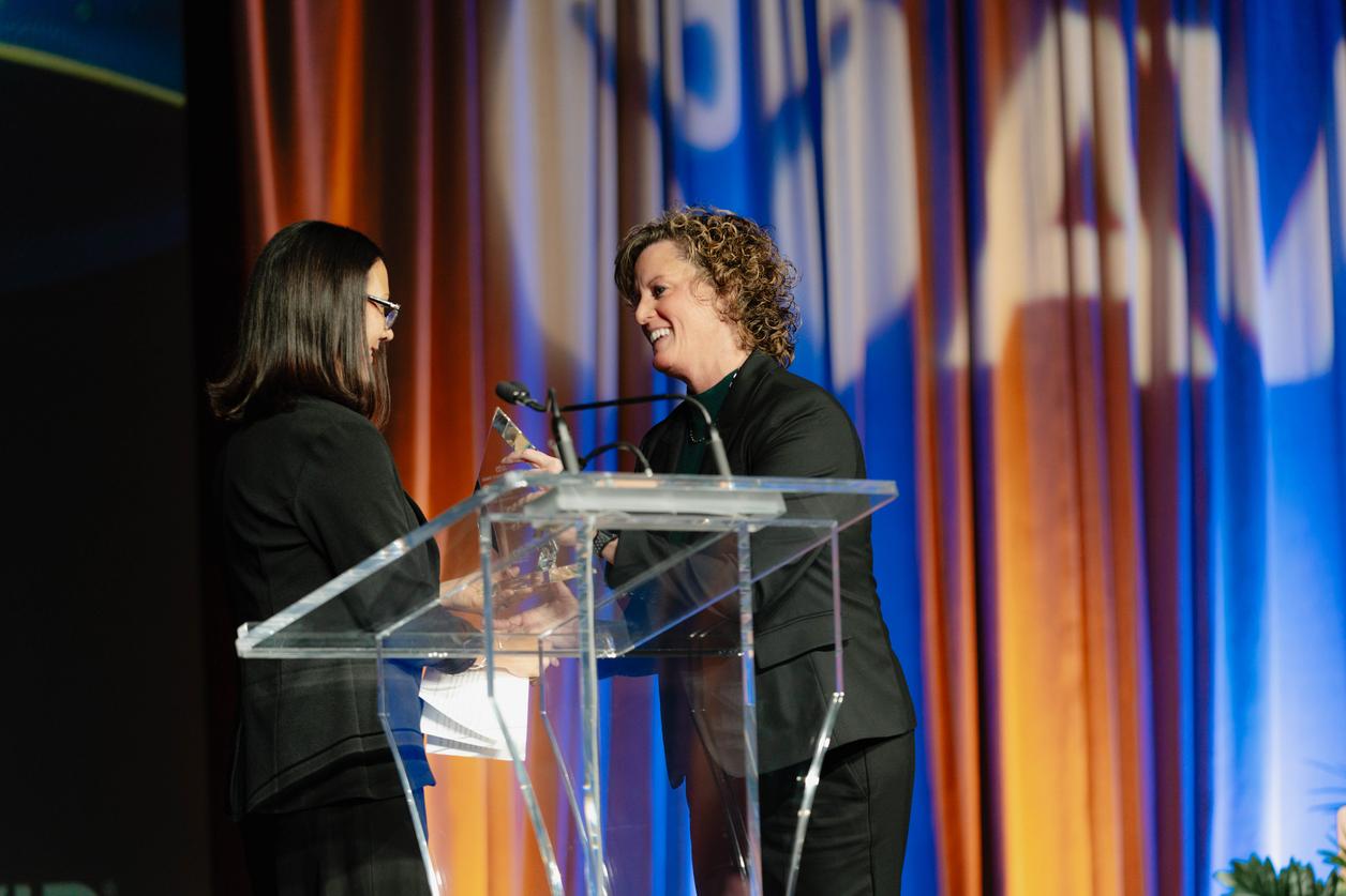 Two individuals standing at a podium on a stage with colorful curtains in the background, engaging in conversation. Two individuals standing at a podium on a stage with colorful curtains in the background, engaging in conversation.