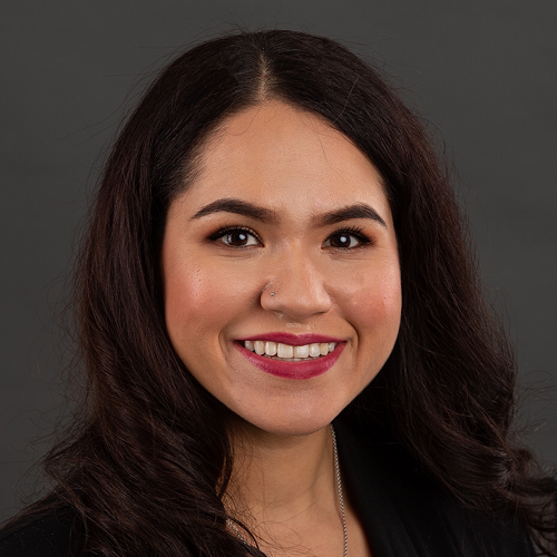 Photograph of a person with long, curly dark hair, wearing a black top against a dark background. The face is censored.