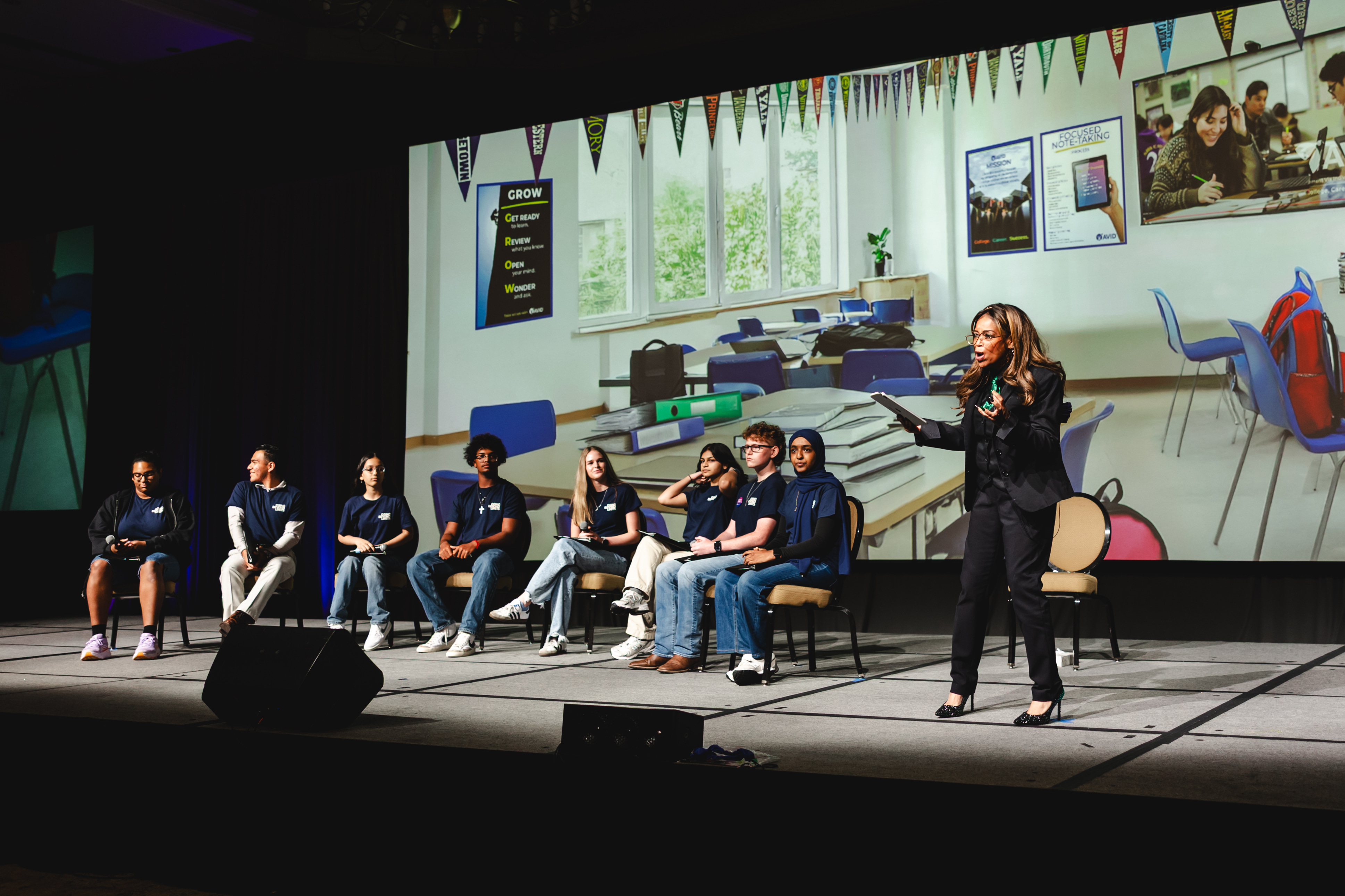 A group of individuals sitting on chairs on a stage with a woman standing and speaking, a classroom background displayed on the screen behind them.