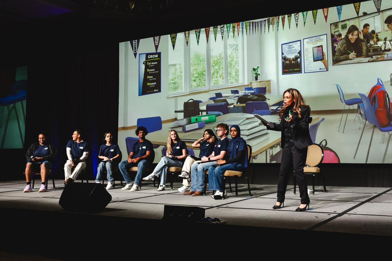 A group of individuals sitting on chairs on a stage with a woman standing and speaking, a classroom background displayed on the screen behind them. A group of individuals sitting on chairs on a stage with a woman standing and speaking, a classroom background displayed on the screen behind them.