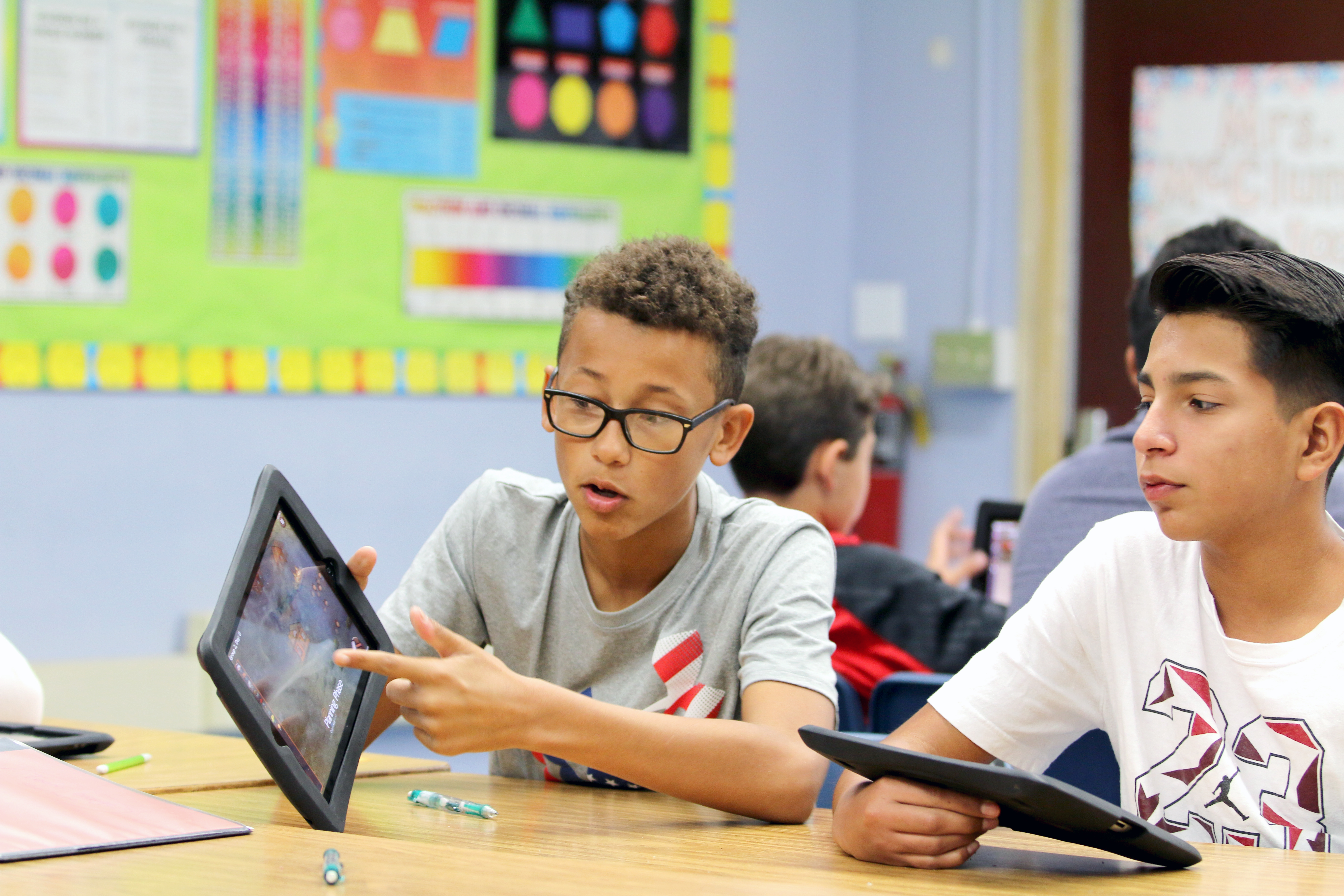 Students in a classroom setting working on tablets, with educational posters and charts on the wall in the background.