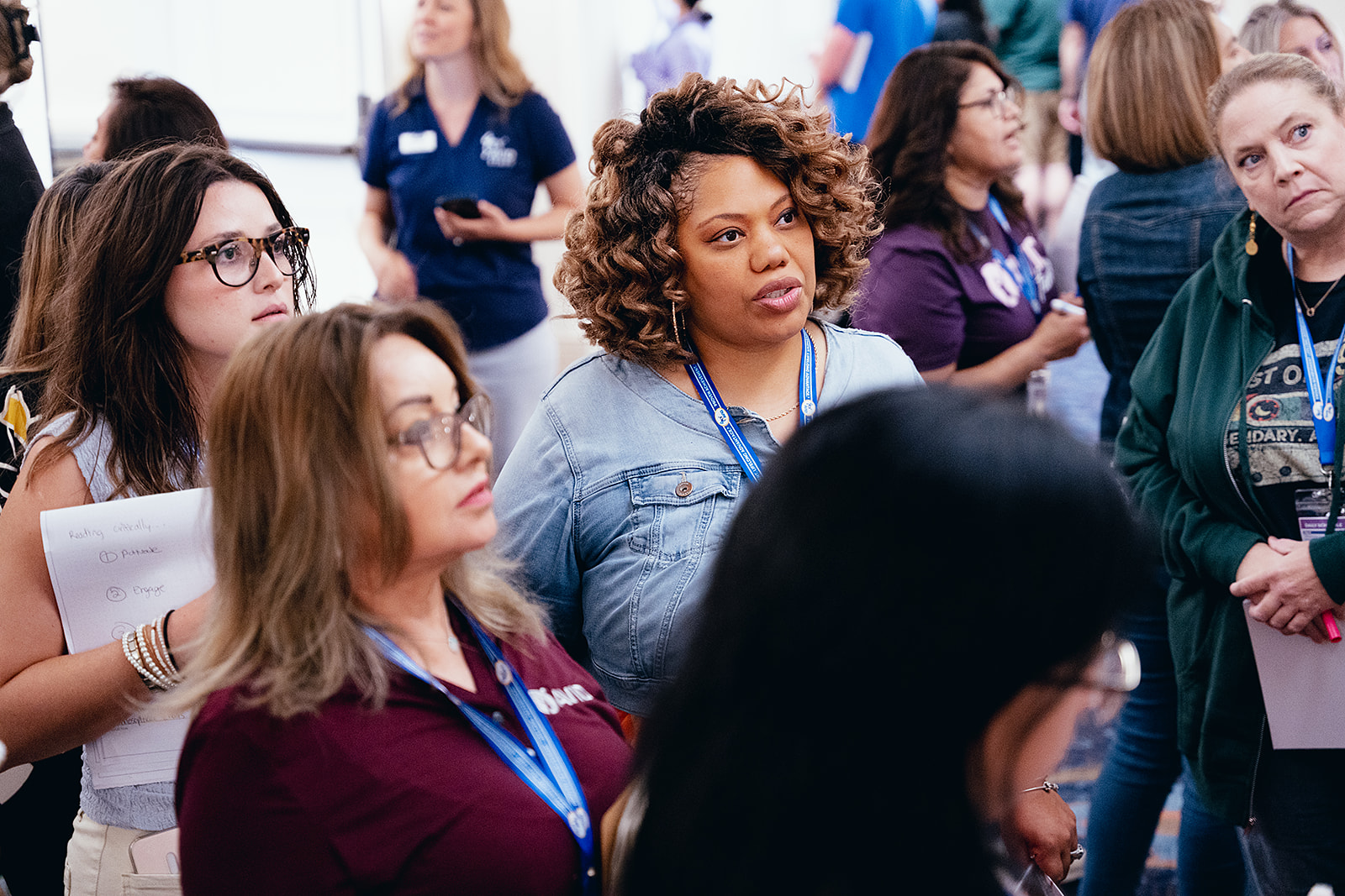 A group of people engaged in a conversation at an indoor event, with some participants holding notebooks and wearing lanyards.