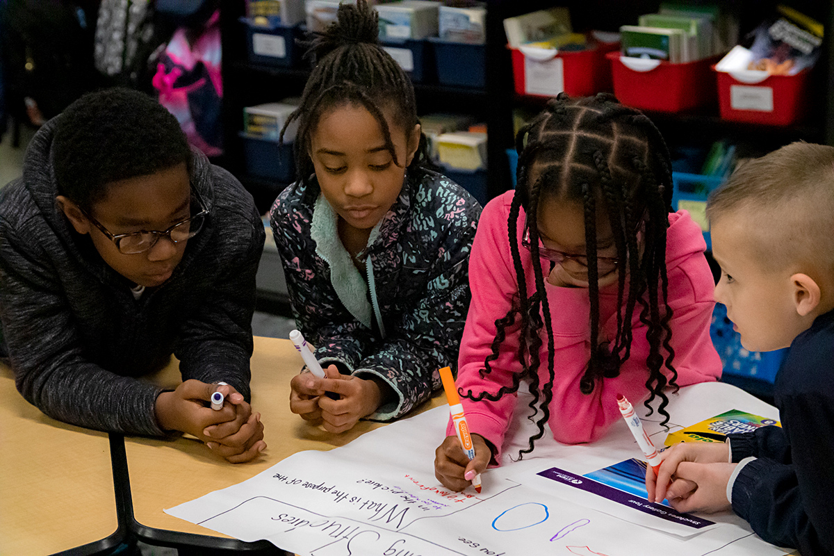 Children working together at a table on a colorful poster with markers in a classroom setting.
