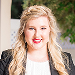 Portrait of a person with long, wavy, blonde hair wearing a dark blazer and white blouse.