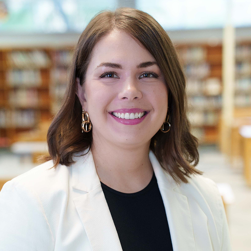 Person wearing a white blazer and black shirt, standing in a library with blurred bookshelves in the background.