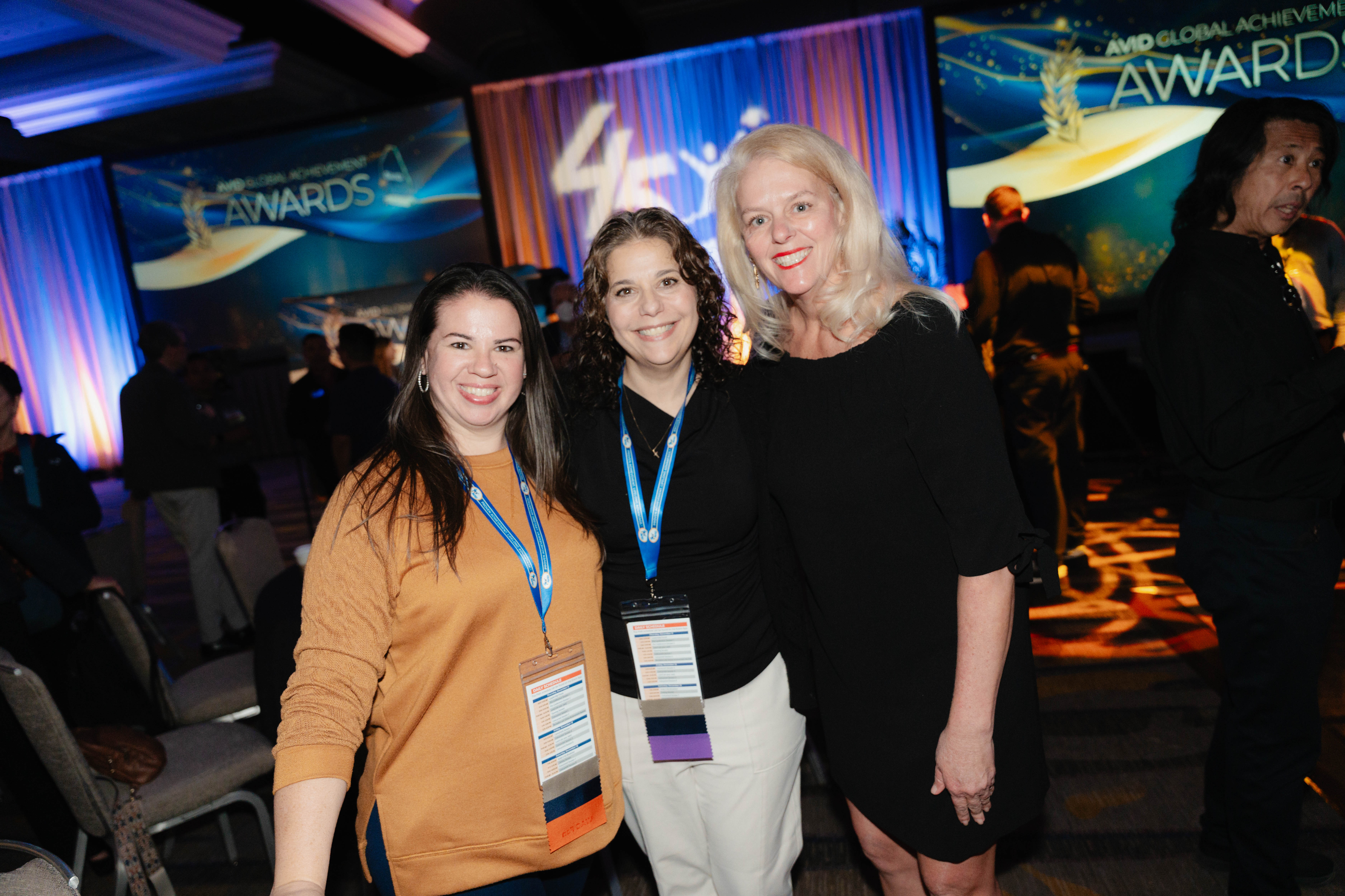Three individuals standing together at an awards ceremony with a staged backdrop and attendees in the background.