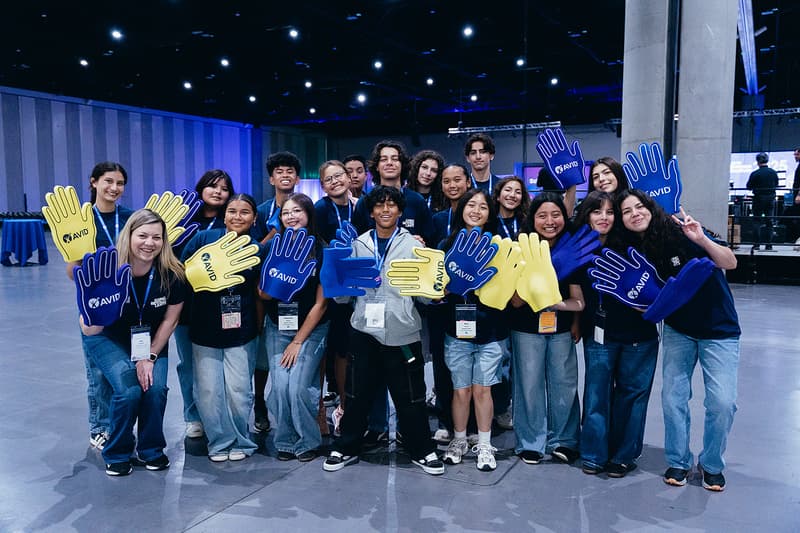 Group photo of people in a large room holding blue and yellow foam hands with 'AVID' written on them. Group photo of people in a large room holding blue and yellow foam hands with 'AVID' written on them.