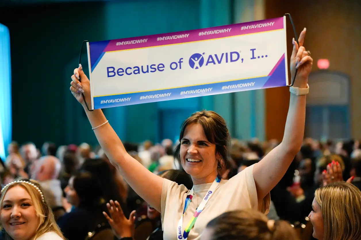 Person smiling and holding a sign that reads 'Because of AVID, I...' in a crowded indoor setting. Person smiling and holding a sign that reads 'Because of AVID, I...' in a crowded indoor setting.