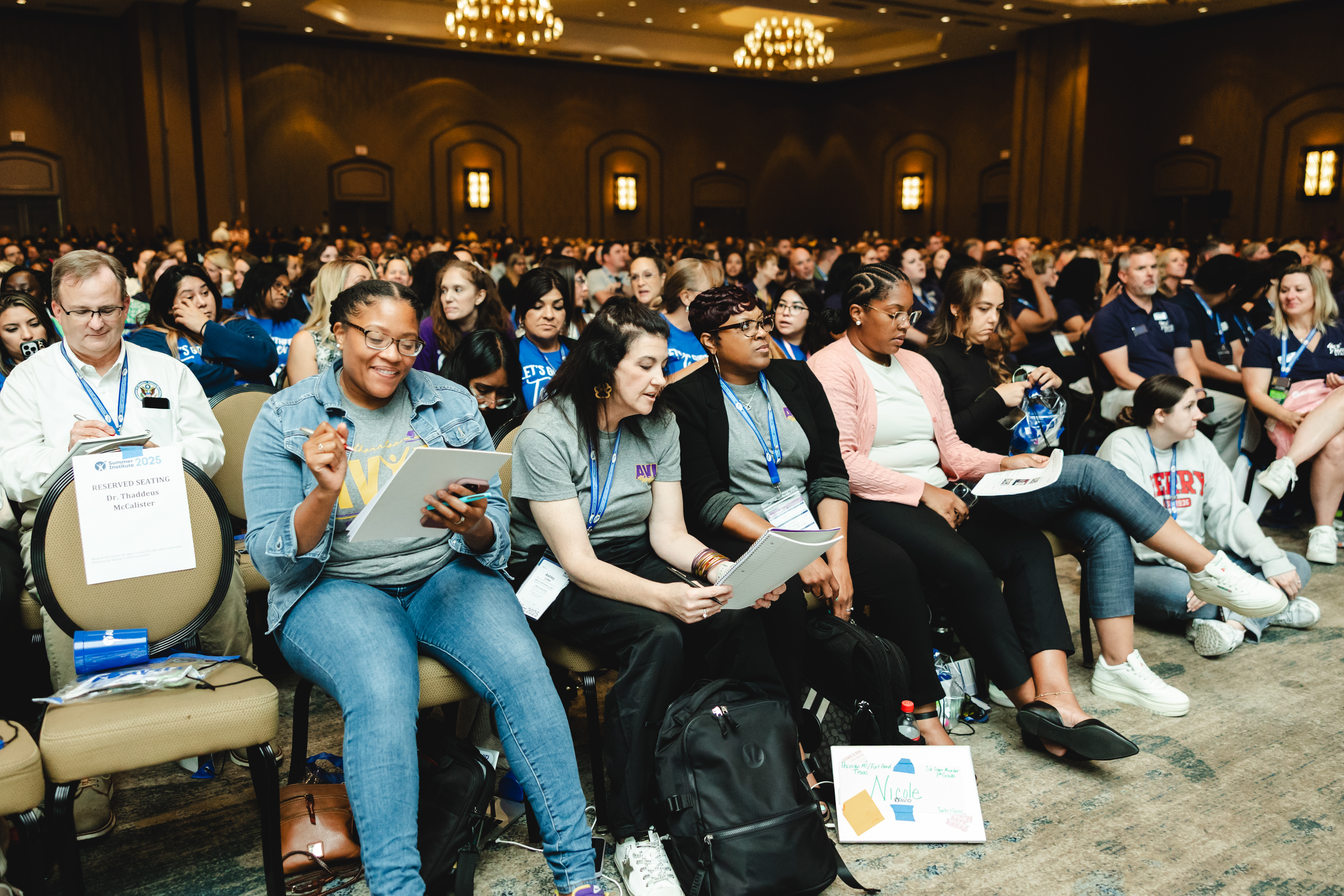 Large group of people seated and attentively reading documents at a conference in a well-lit hall with chandeliers.