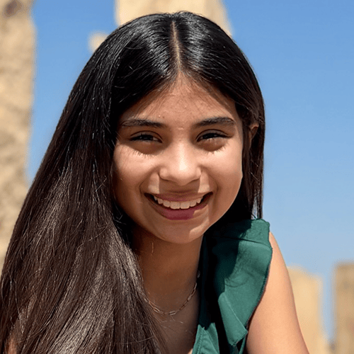 Individual with long, straight dark hair, wearing a green top, sitting outdoors with blurred background. Individual with long, straight dark hair, wearing a green top, sitting outdoors with blurred background.