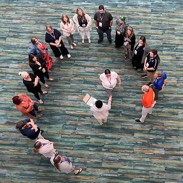 A group of people standing in a circle in an indoor setting with a blue-green floor. A group of people standing in a circle in an indoor setting with a blue-green floor.