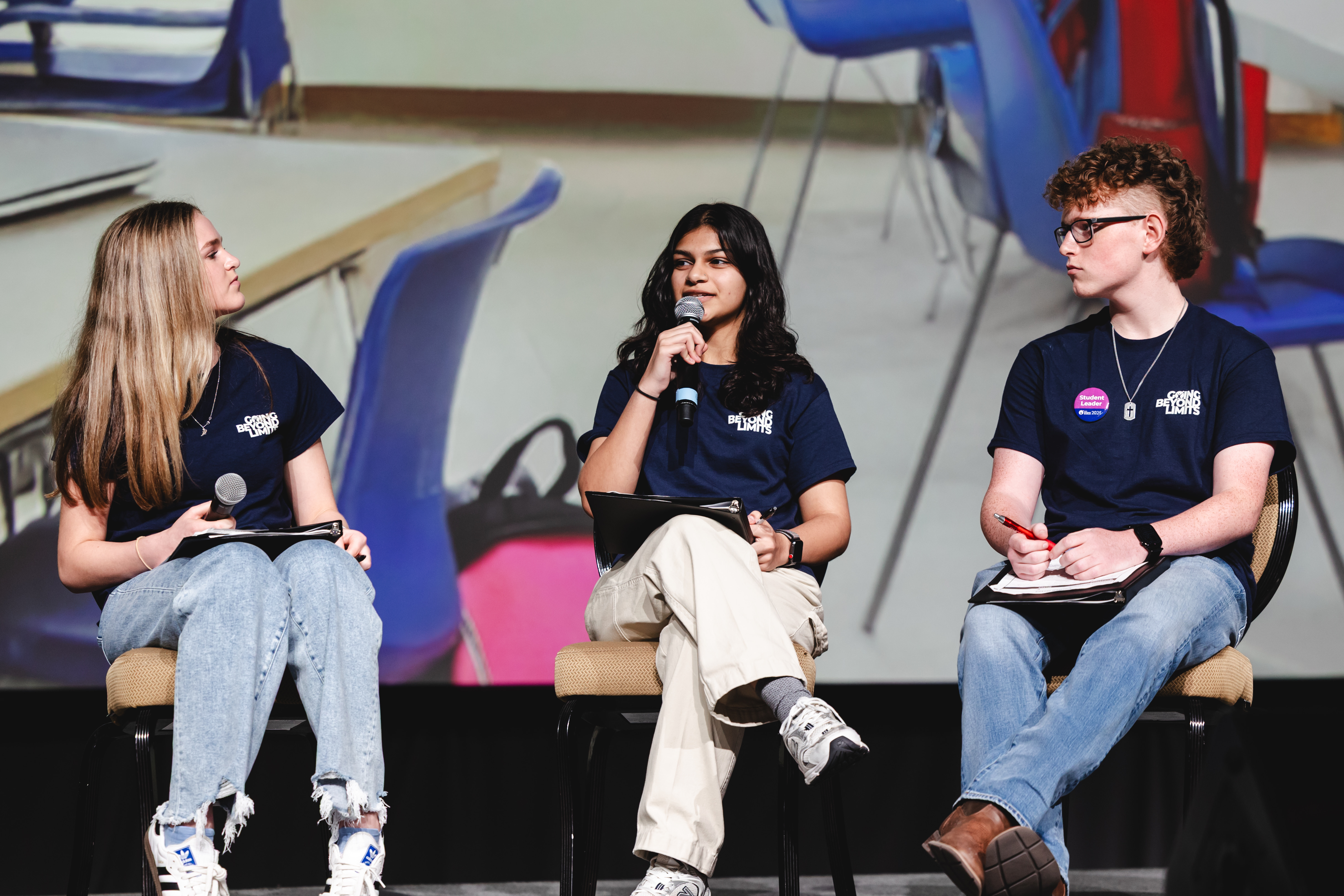 Three individuals sitting on chairs engaging in a discussion, each holding a microphone and wearing blue shirts, with a classroom-like background.
