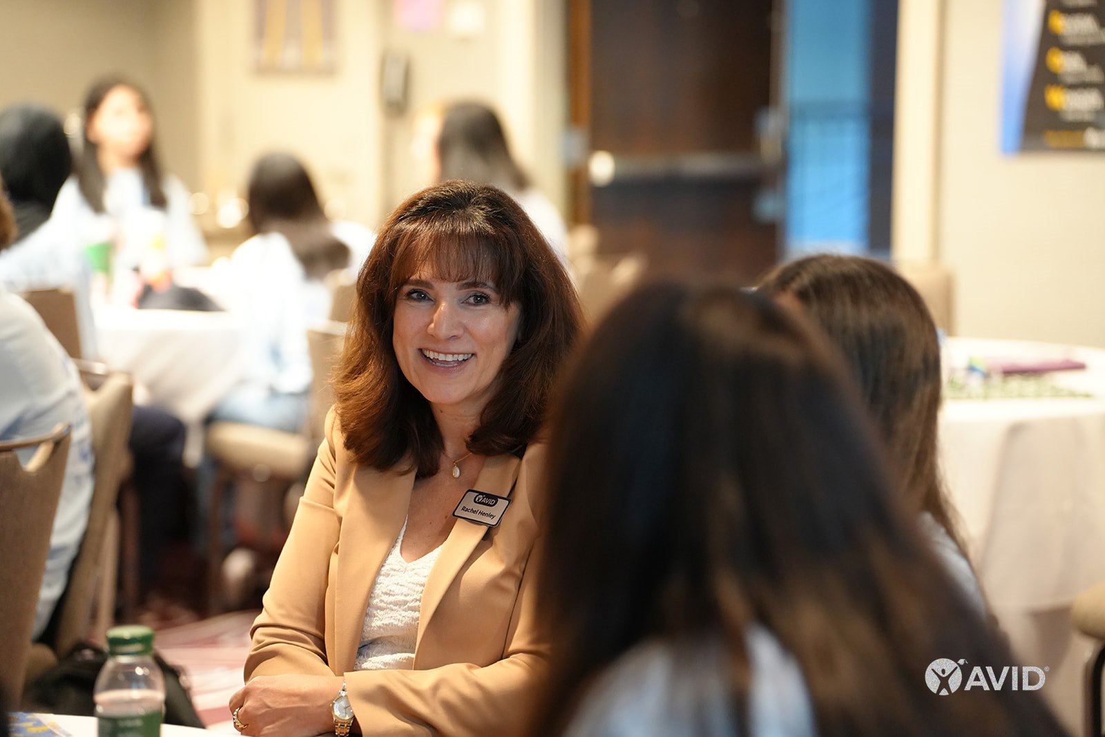A person sitting at a table during an event, smiling, and looking at the camera. The background has several people seated and blurred out.