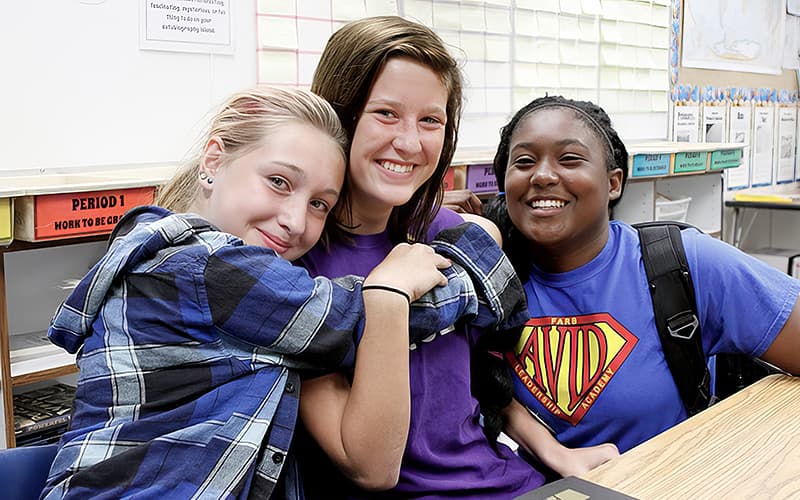 Three people in a classroom, one person is putting their arm around another. Three people in a classroom, one person is putting their arm around another.