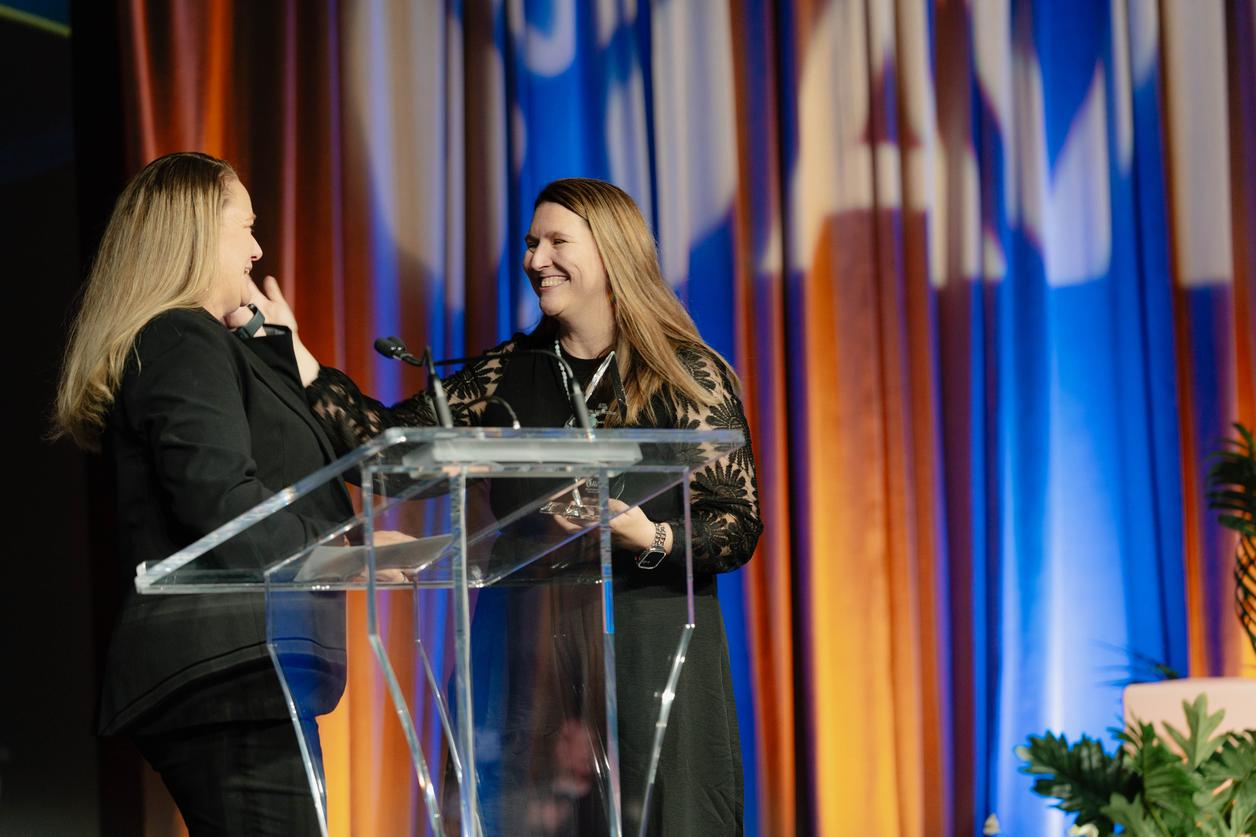 Two people standing behind a podium with microphones, in front of a colorful backdrop Two people standing behind a podium with microphones, in front of a colorful backdrop