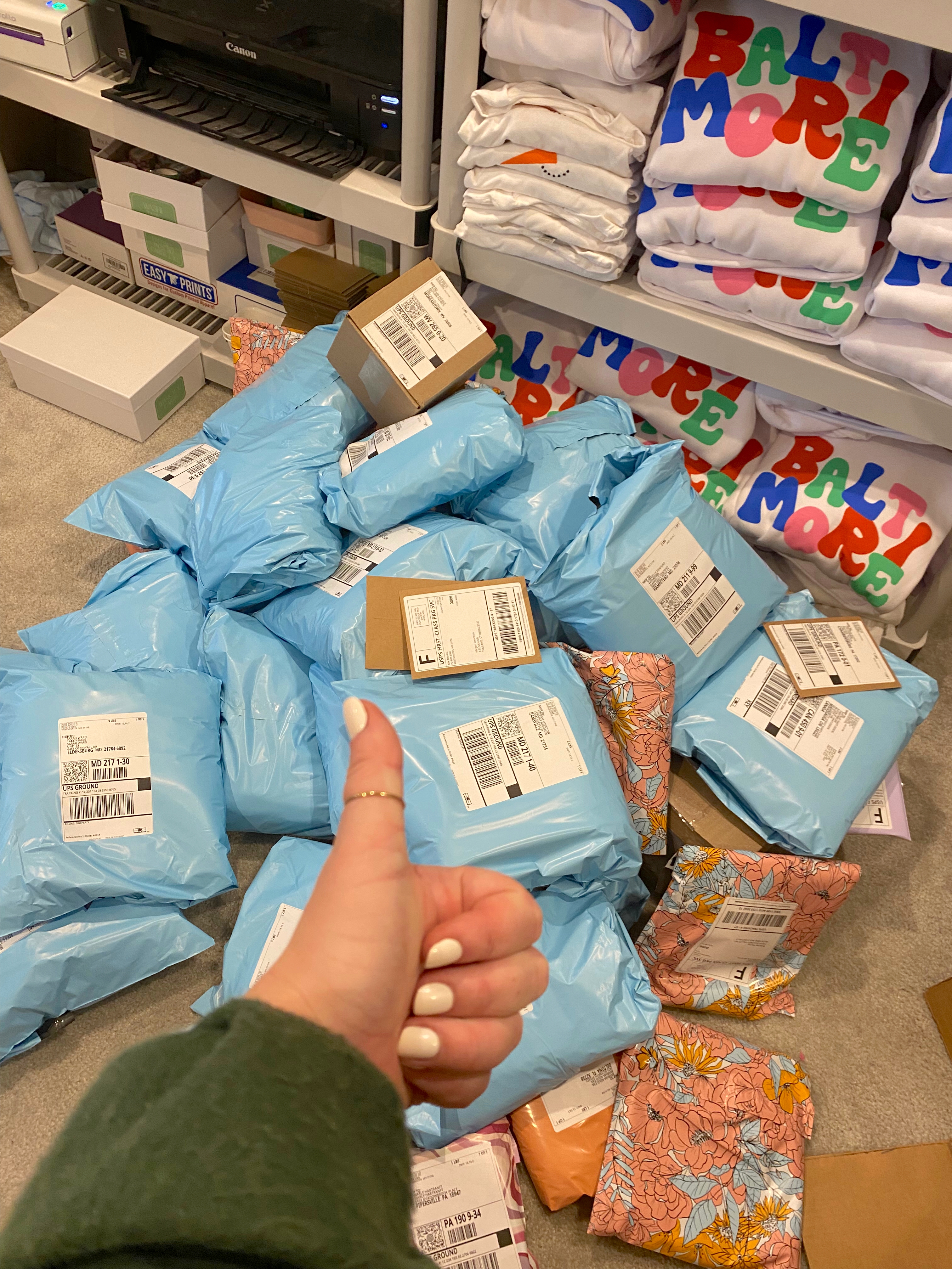 Thumbs up in the foreground with a stack of folded white sweaters with Baltimore printed in multiple colors and blue mail bags in the background.