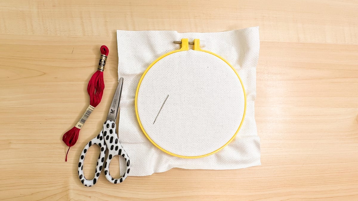 Embroidery materials laid out on the table including fabric, embroidery hoop, need, thread, and scissors.