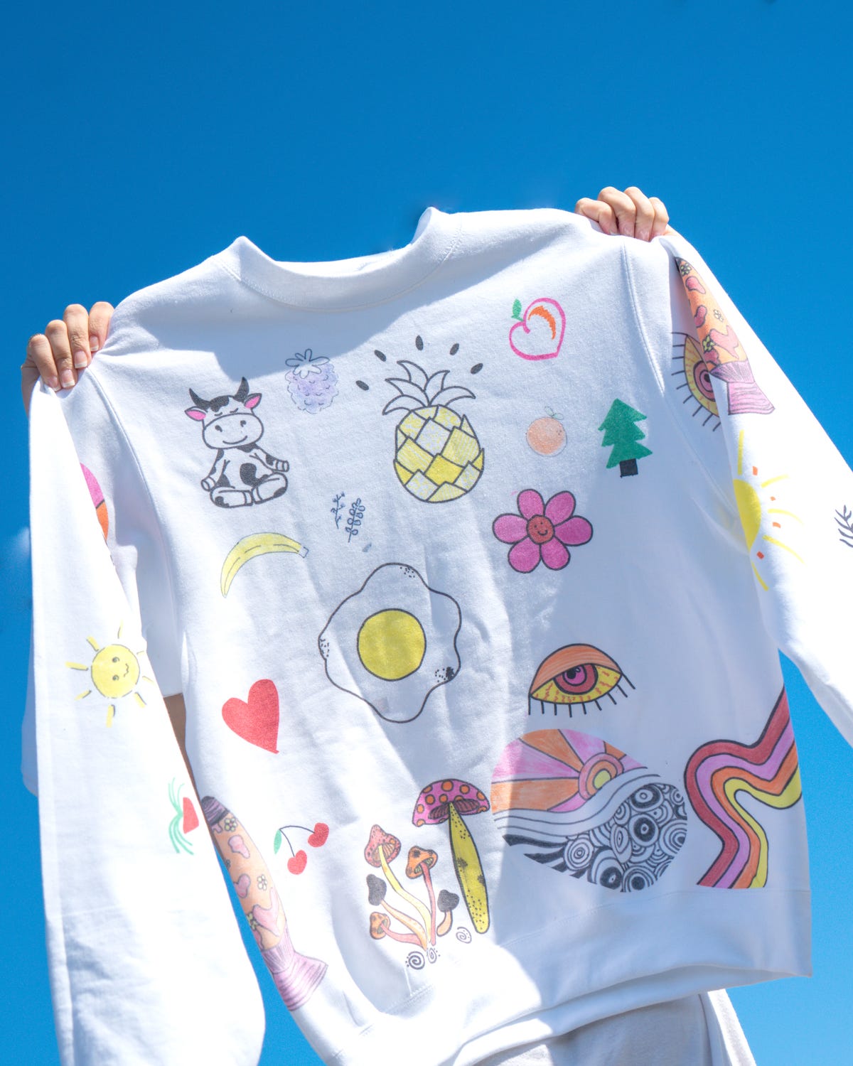 Two hands holding up a white crewneck sweatshirt with doodles on the front.