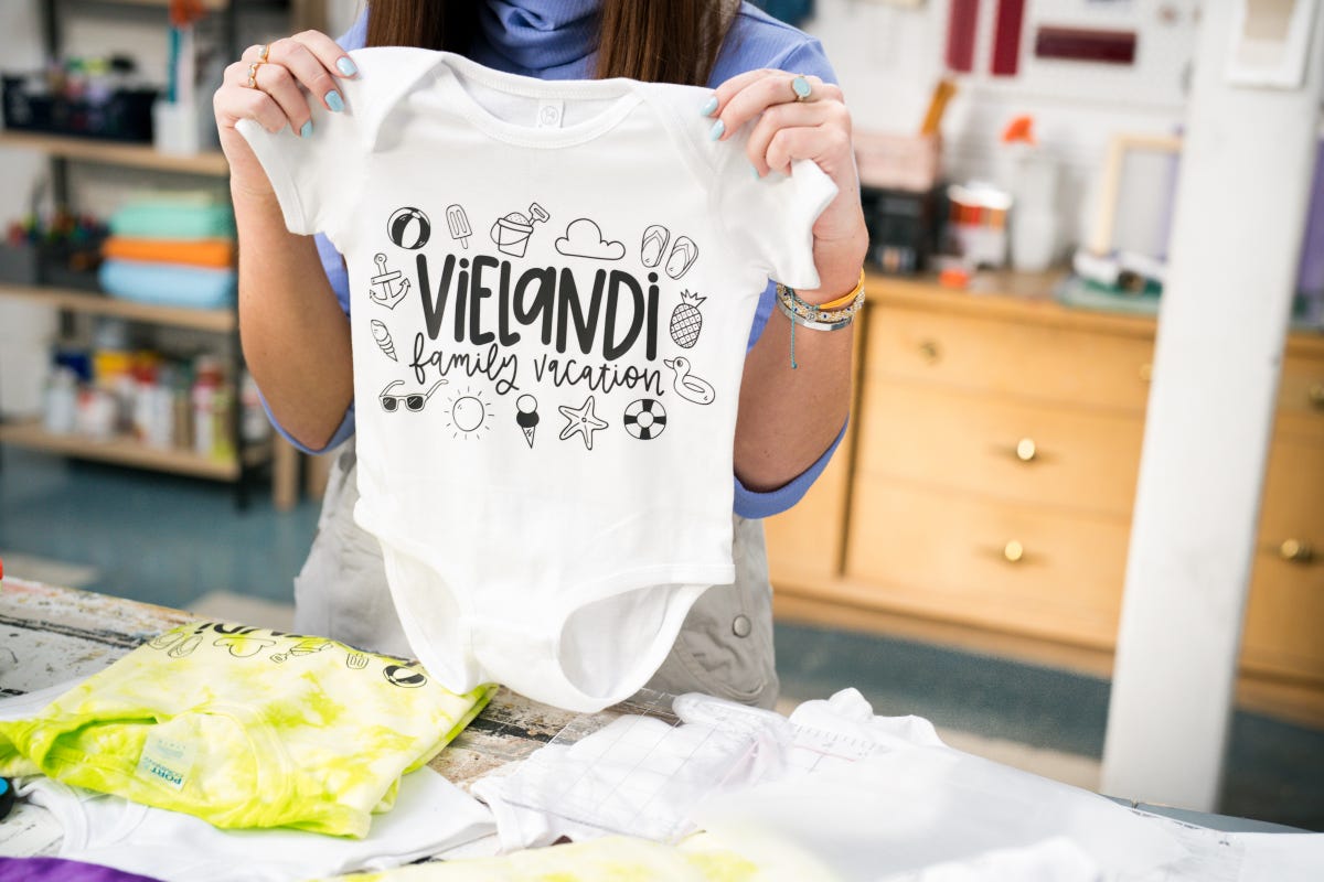 Allie holding up the Rabbit Skins Infant Short Sleeve Baby Rib Bodysuit in white pressed with Vielandi Family Vacation in the front.
