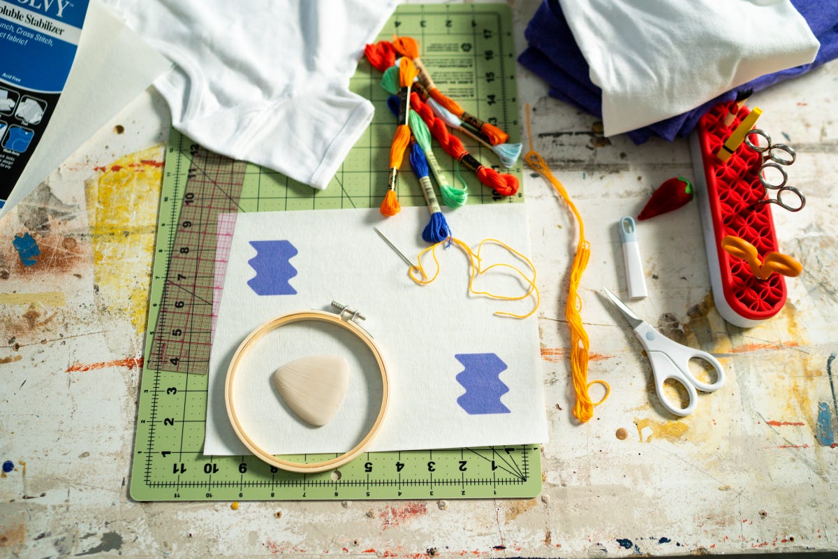 Embroidery tools laid out on a workspace including embroidery hoop, threads, stabilizer with printed design, and scissors.