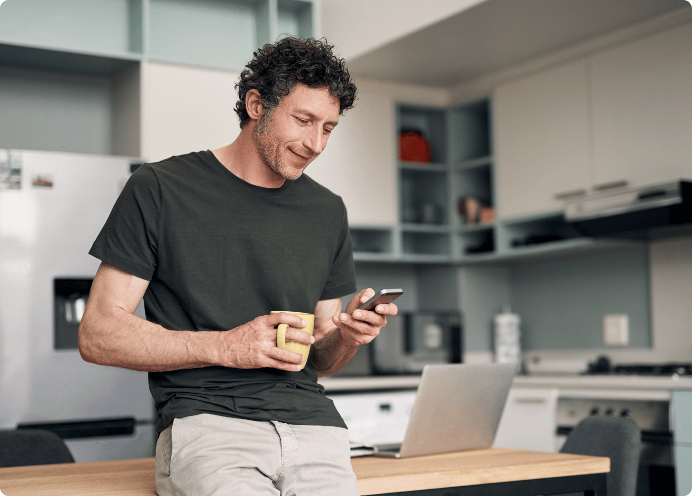 Man sitting on kitchen counter with phone in hand and laptop in background