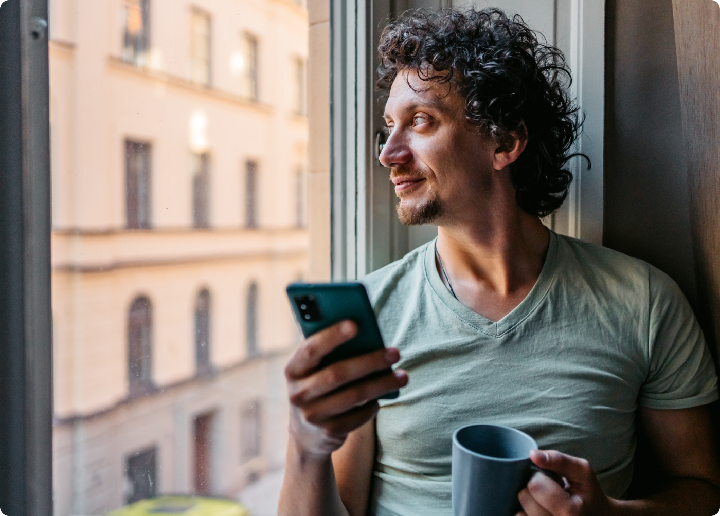 Man looking out his window with smartphone and coffee mug in hand
