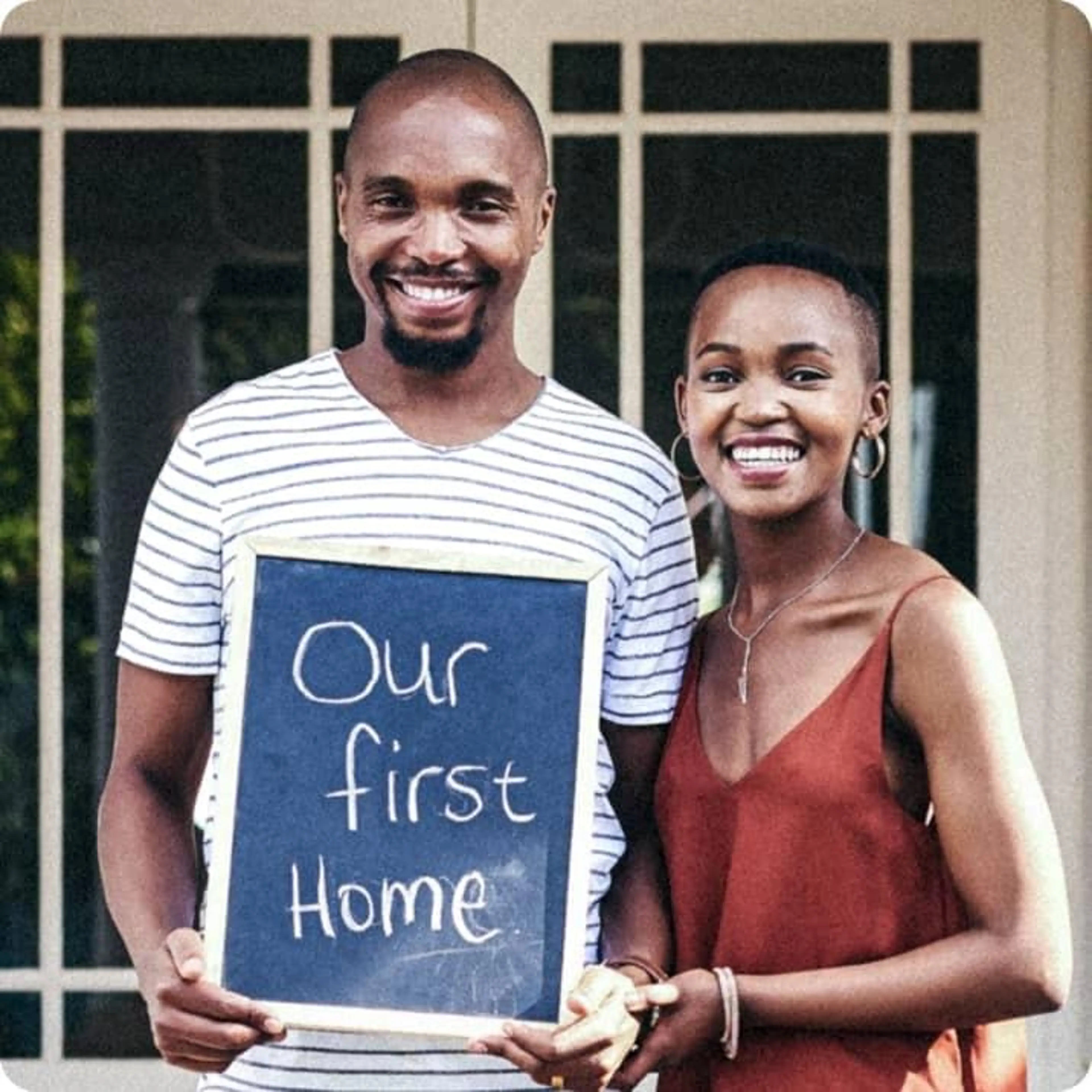 A couple smiling while holding a sign that says "our first home".