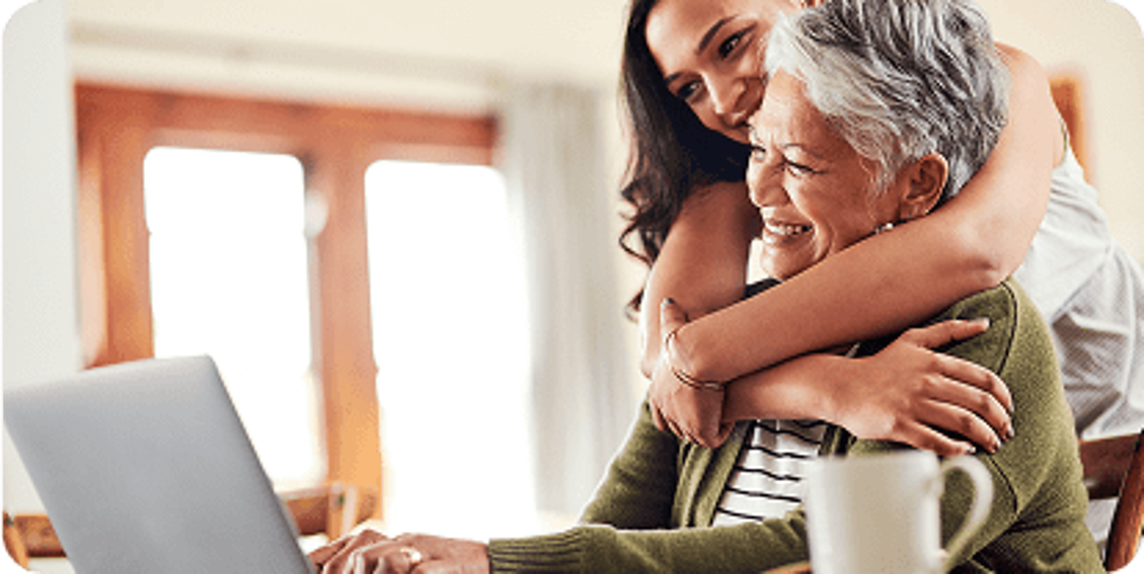 Woman wrapping her arms around an older woman as they smile looking at a laptop screen.