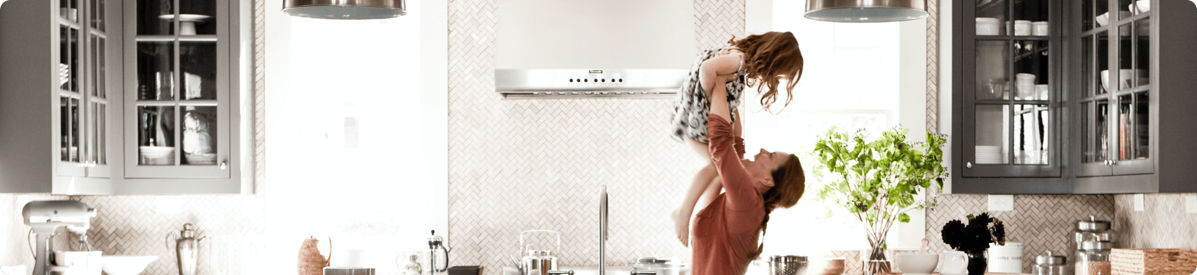 A mother lifts her young child up in the air inside a bright, modern kitchen.