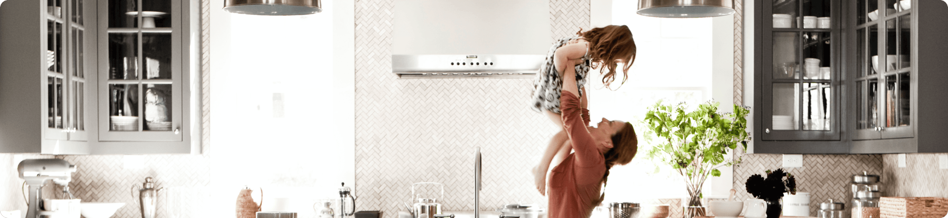 A mother lifts her young child up in the air inside a bright, modern kitchen.