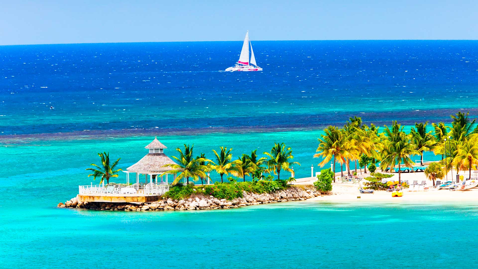 Ocho Rios beach in Jamaica, with white sand, palm trees, turquoise Caribbean Sea, and a white sailboat on the horizon.