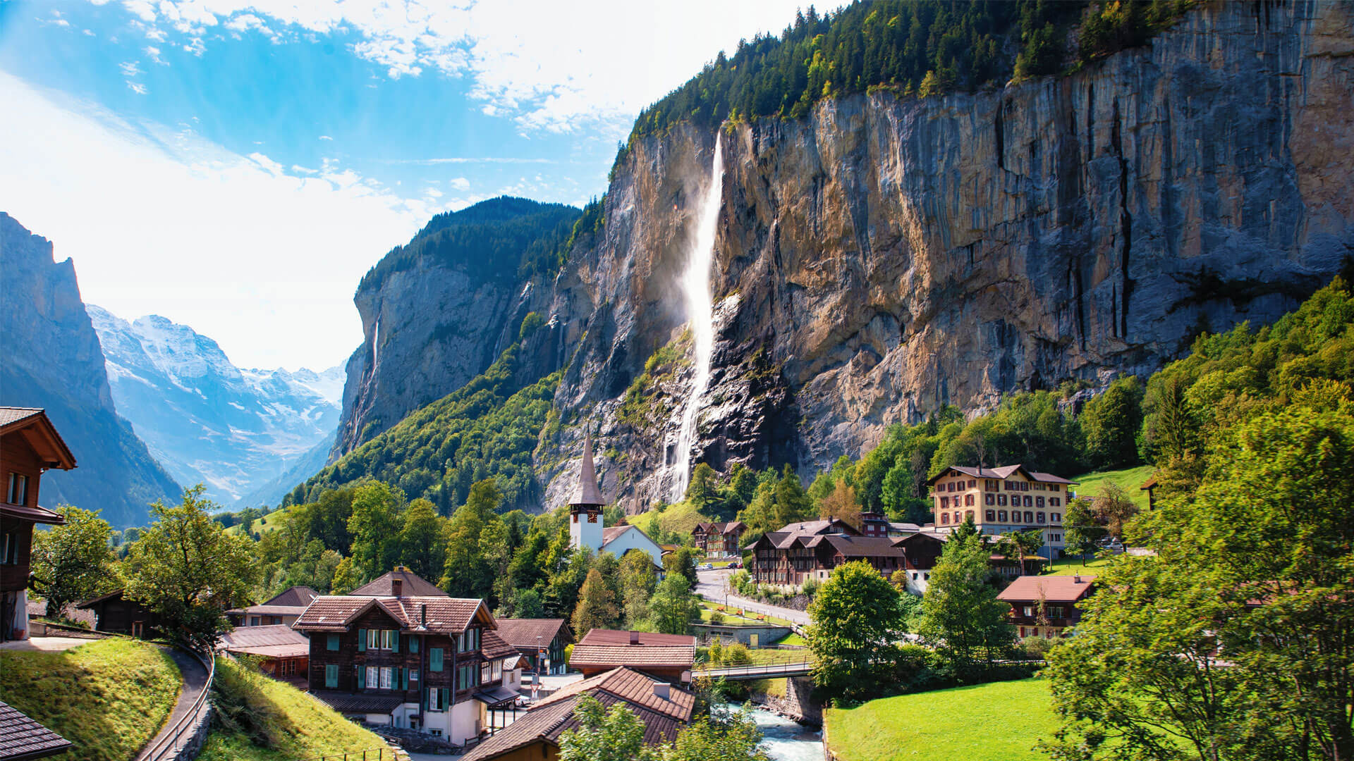 Village suisse alpin avec chalets et cascade tombant d’une falaise dans la vallée de Lauterbrunnen, entourée de montagnes et de verdure.