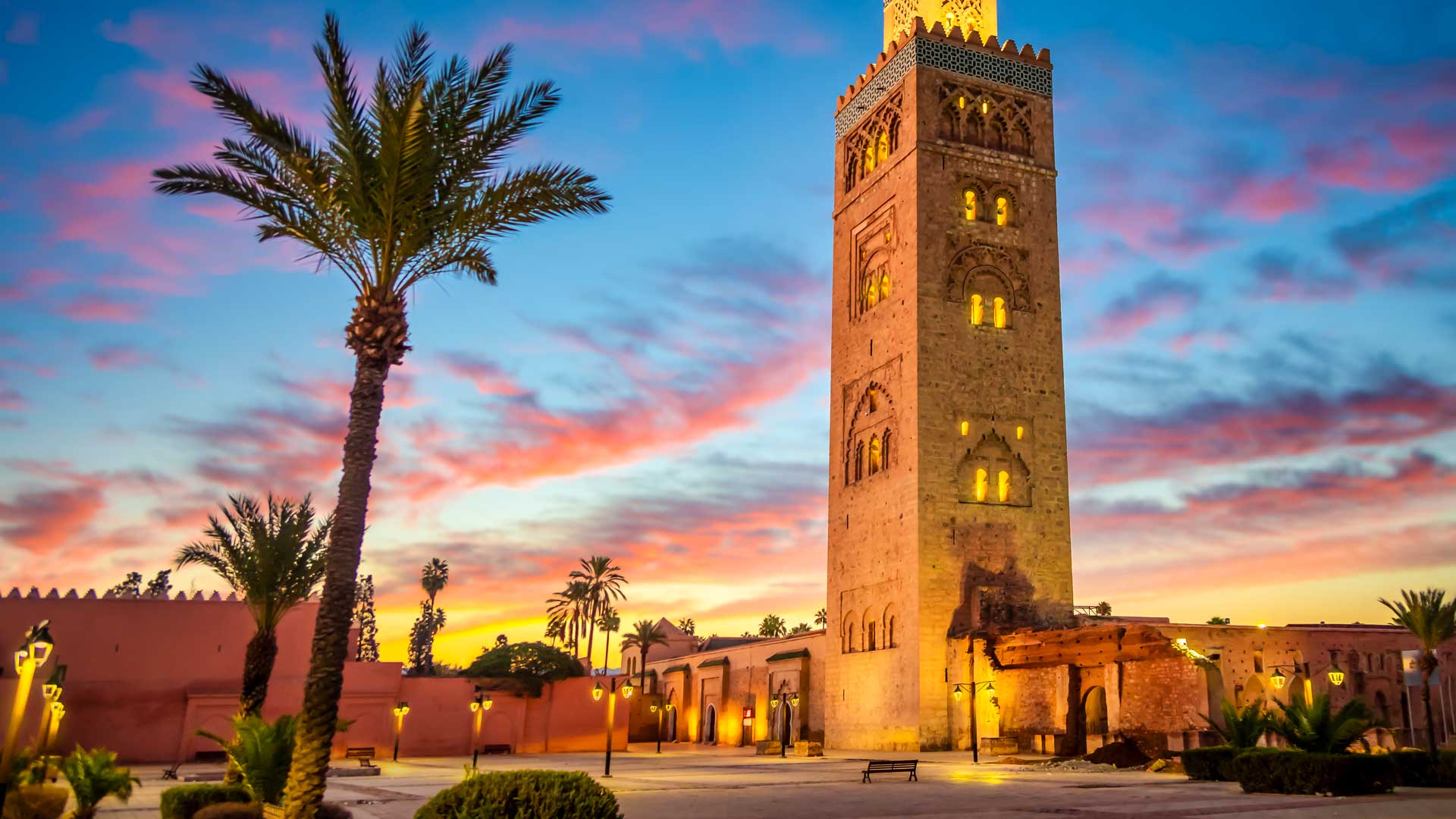 Panoramic view of the Koutoubia Mosque in Marrakech, Morocco, at sunset, with palm trees and a colourful sky.