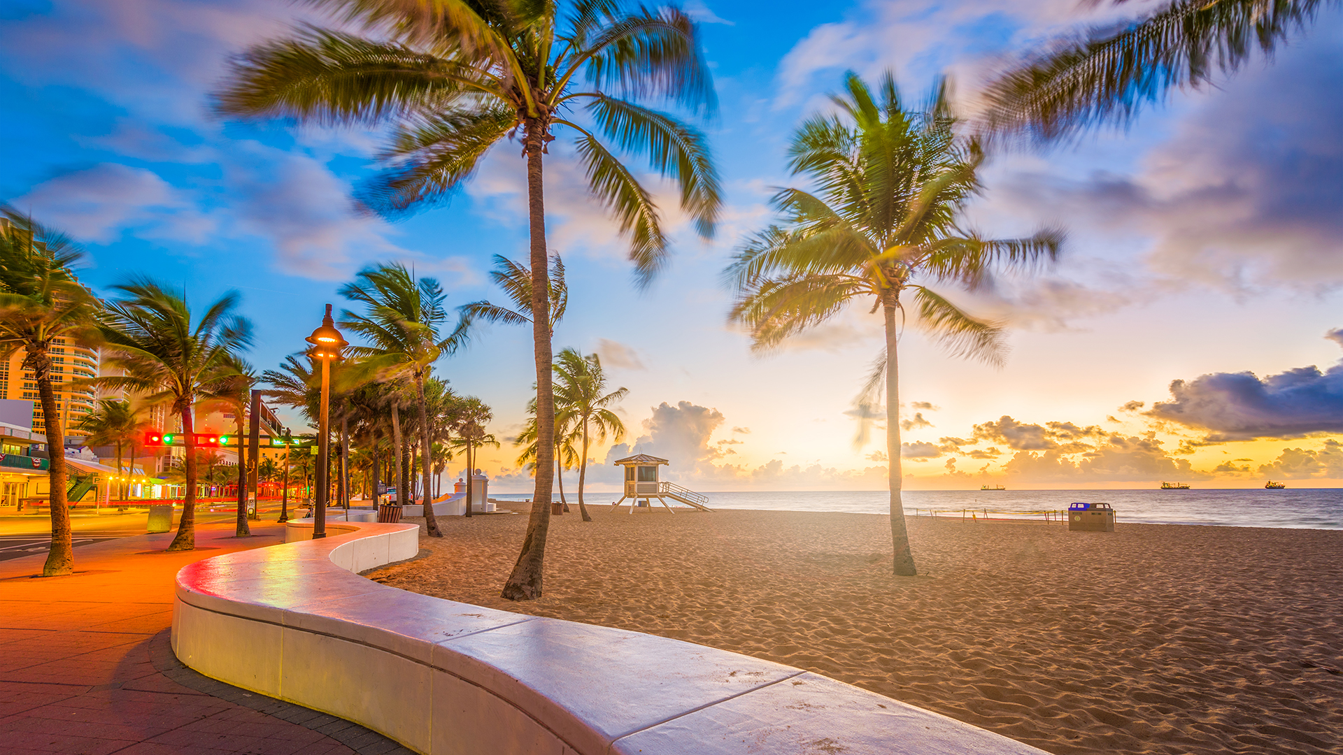 Sunset over the Fort Lauderdale boardwalk in Florida with a sandy beach and palm trees.