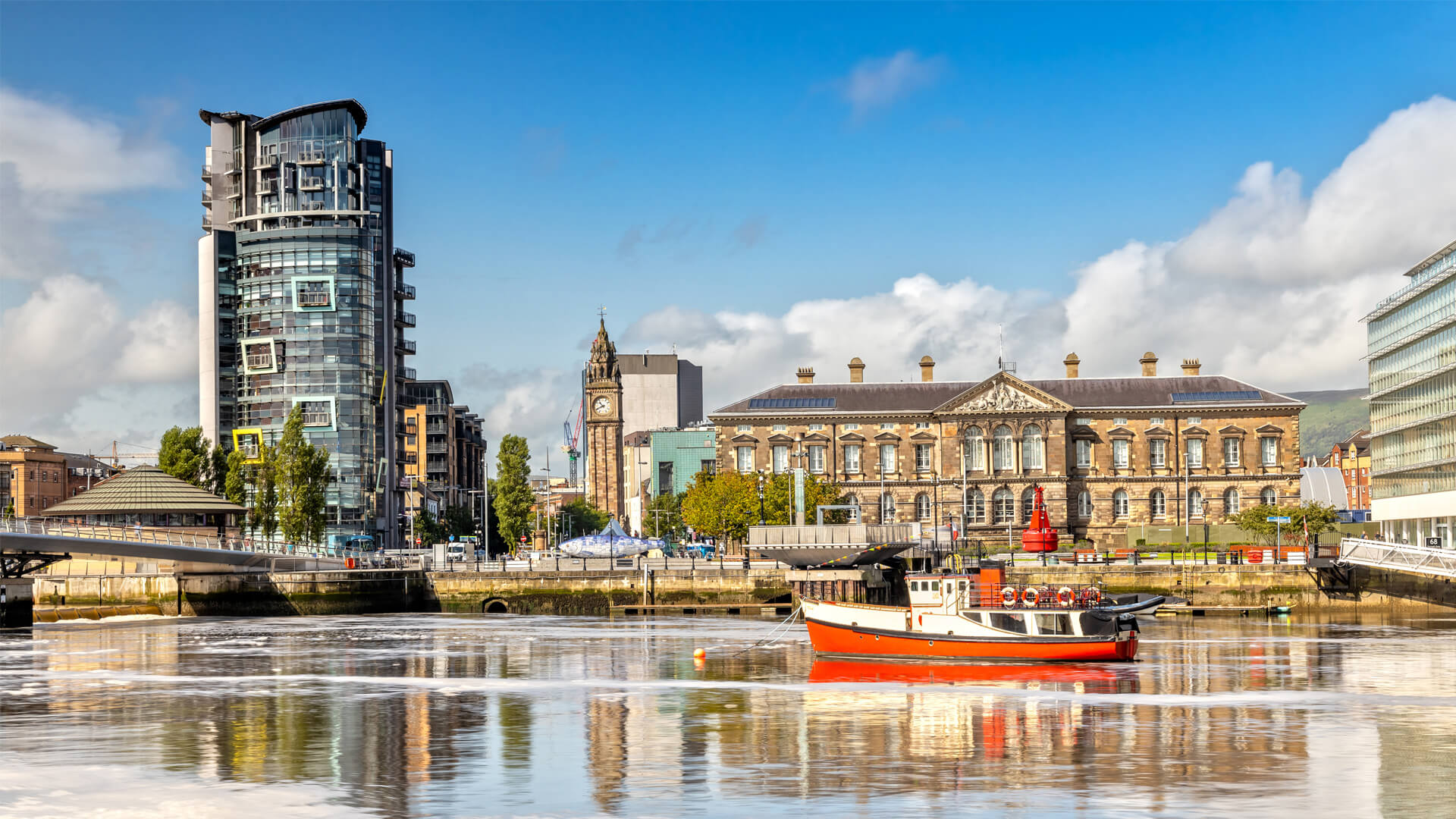 Cityscape of Belfast, Northern Ireland, showing the Victorian Custom House along the River Lagan and a red boat docked nearby.