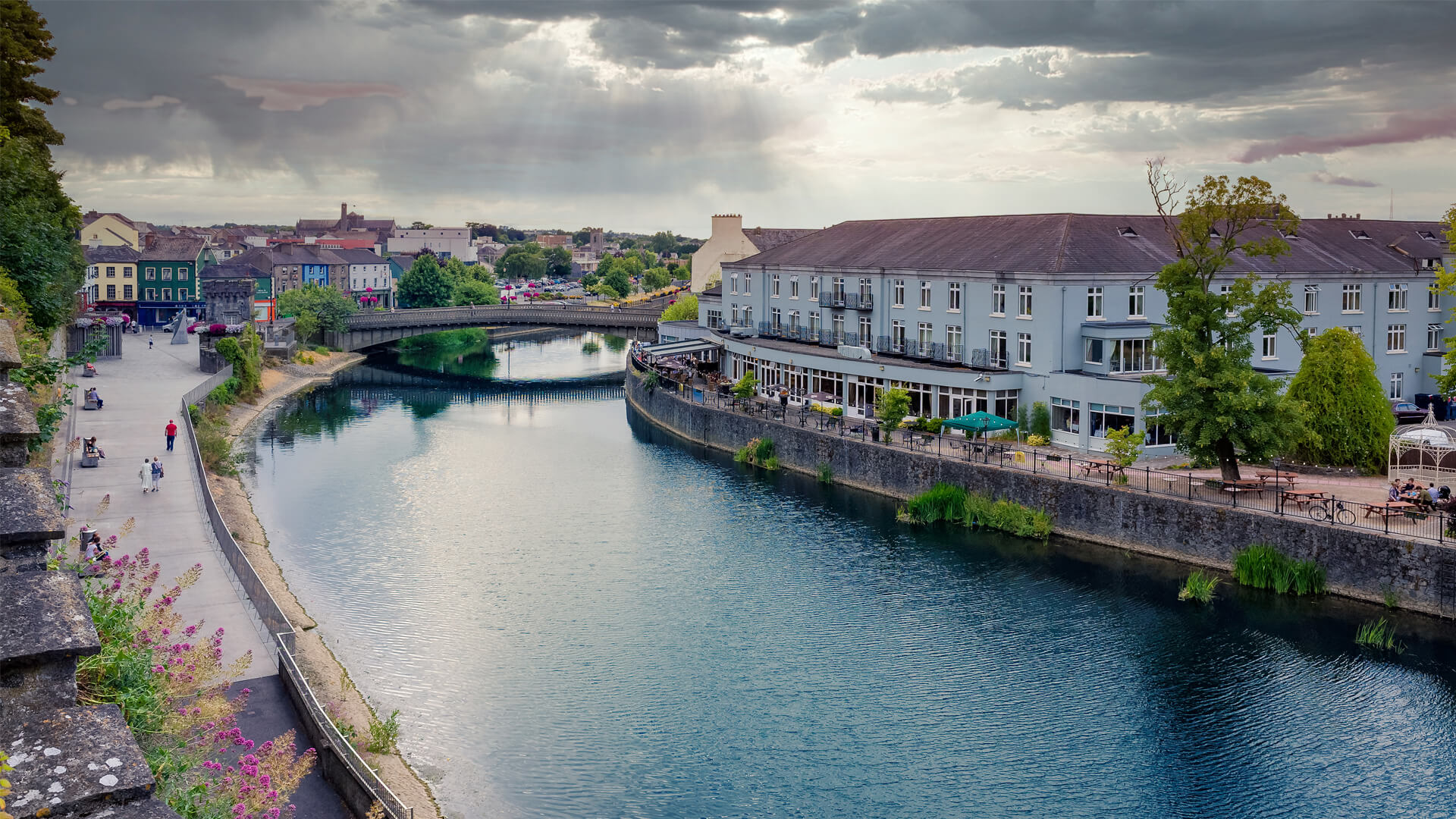 The River Nore in Kilkenny, Ireland, lined with buildings and walkways, crossed by a bridge under a cloudy sky.