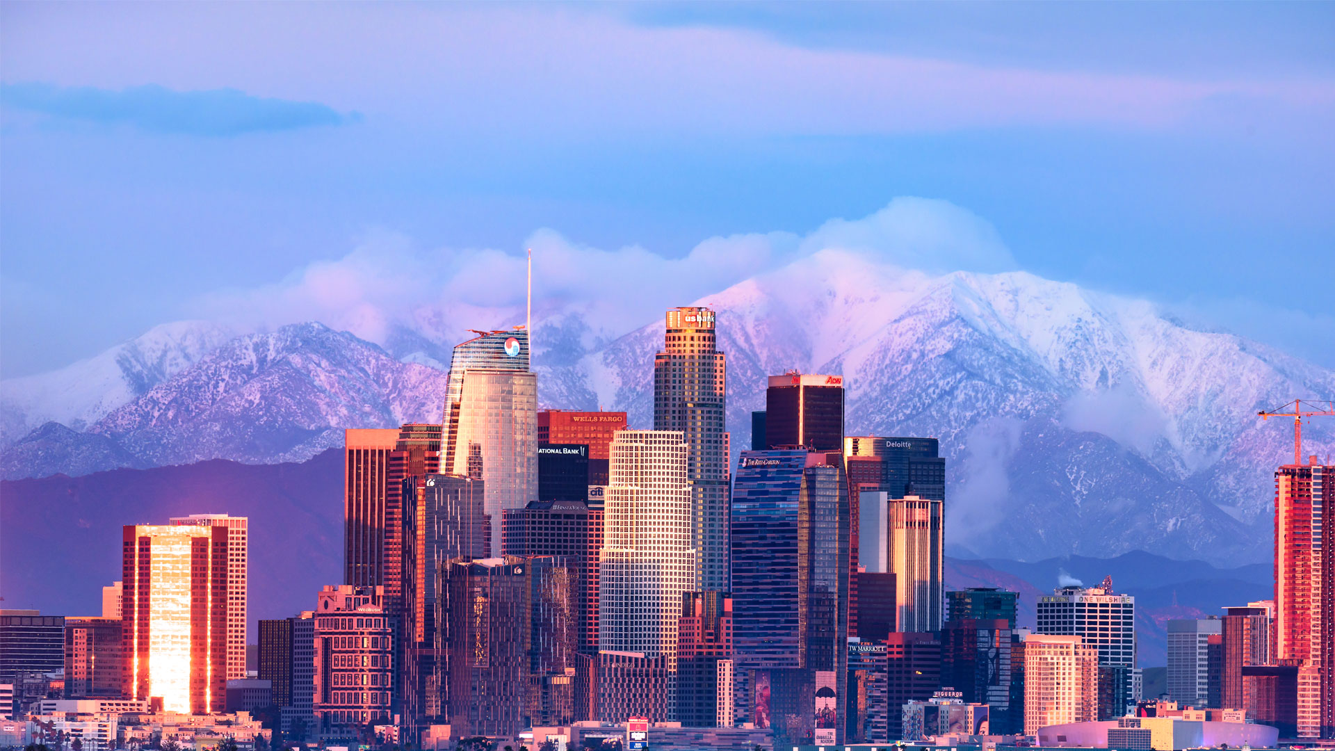 Los Angeles skyscrapers glowing in pink and golden hues at sunset with snow-capped mountains in the background.