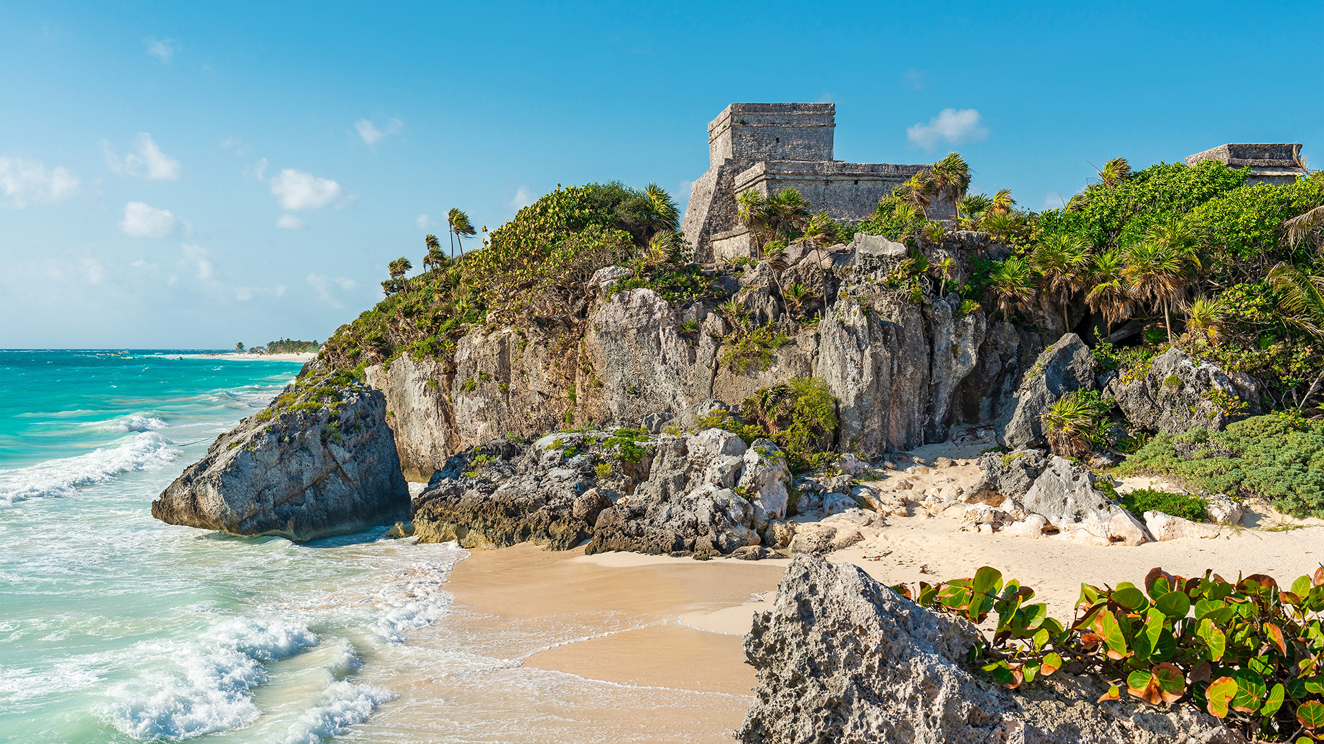 Mayan ruins of Tulum perched on green cliffs overlooking turquoise waters and a white-sand beach. 