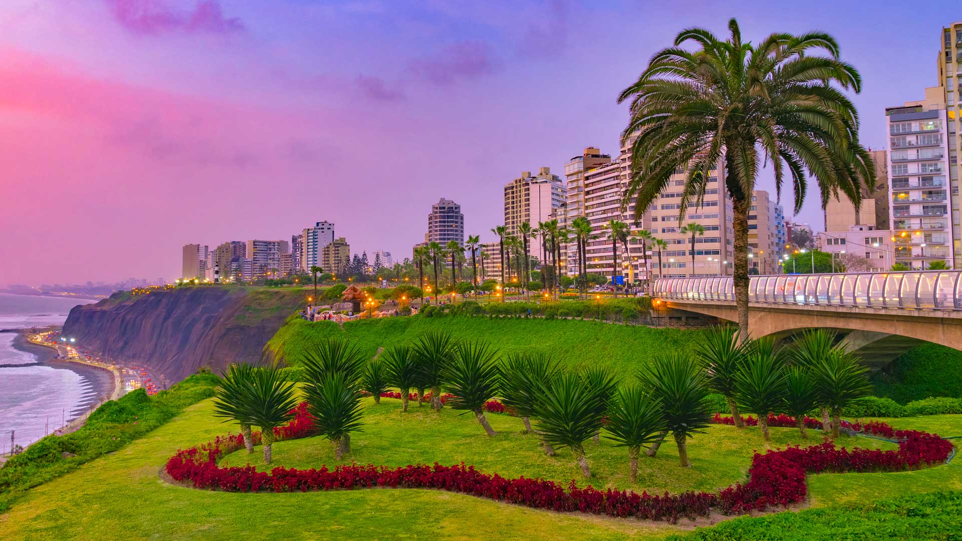 Panoramic view of Lima, Peru, overlooking the Pacific Ocean with buildings and palm trees set against a violet sunset.