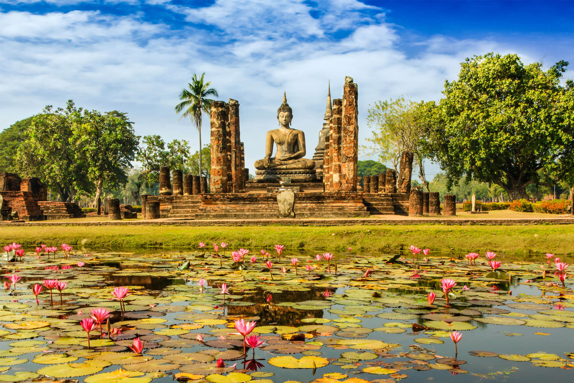 Seated Buddha statue at Sukhothai Historical Park in Thailand, surrounded by ruins and a blooming lotus pond.
