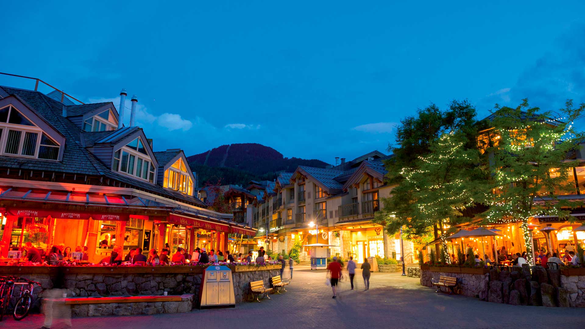Pedestrian village of Whistler at dusk, with illuminated alpine lodges, lively restaurants and mountains in the background.