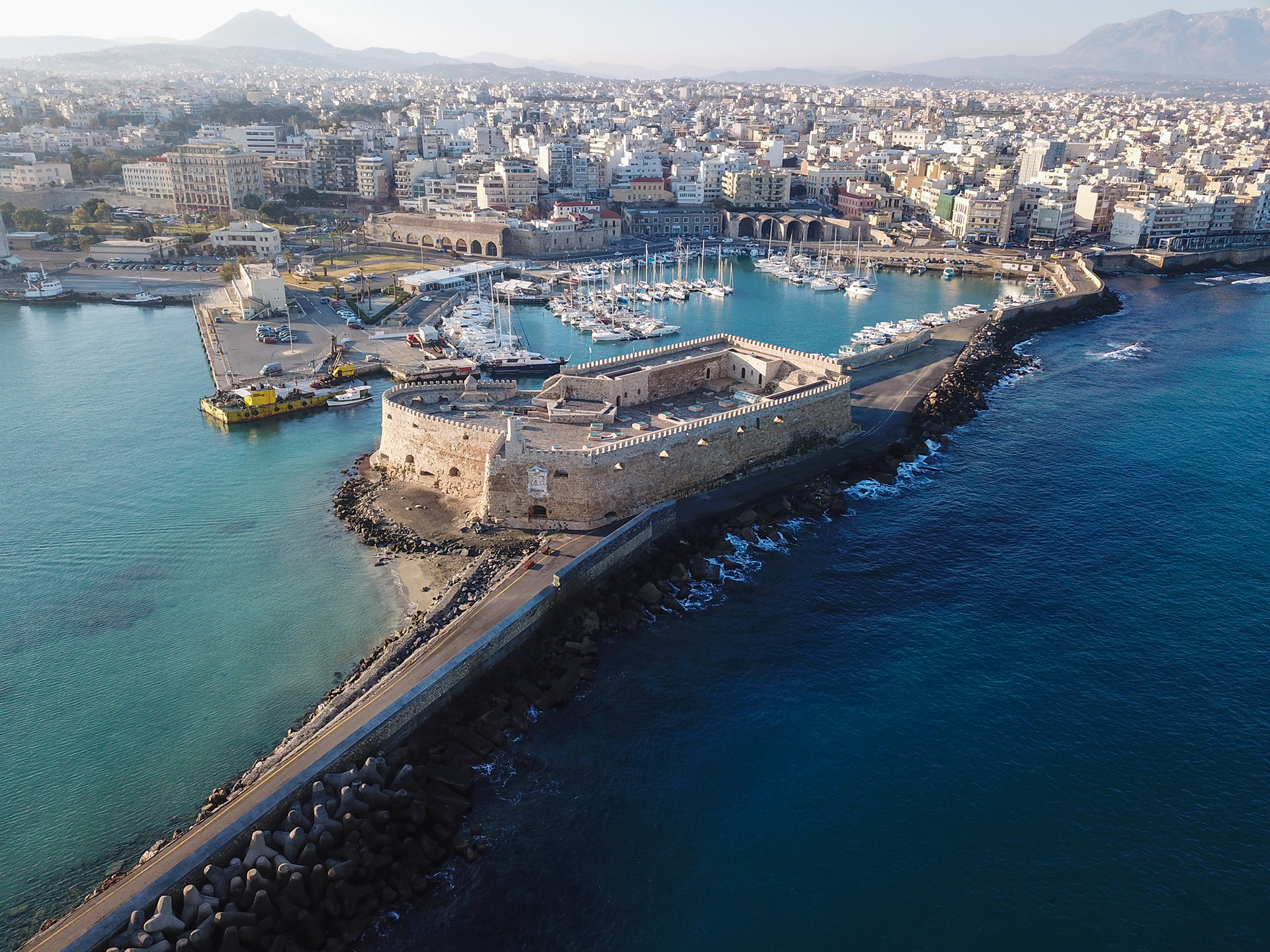 Vue aérienne du port d’Héraklion en Crète, avec sa forteresse vénitienne entourée par la mer Égée et la ville en arrière-plan.