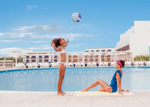 Près d’une piscine dans un Club Med des Caraïbes, une mère assise aux abords d’une piscine regarde sa fille jouer avec un ballon de plage.