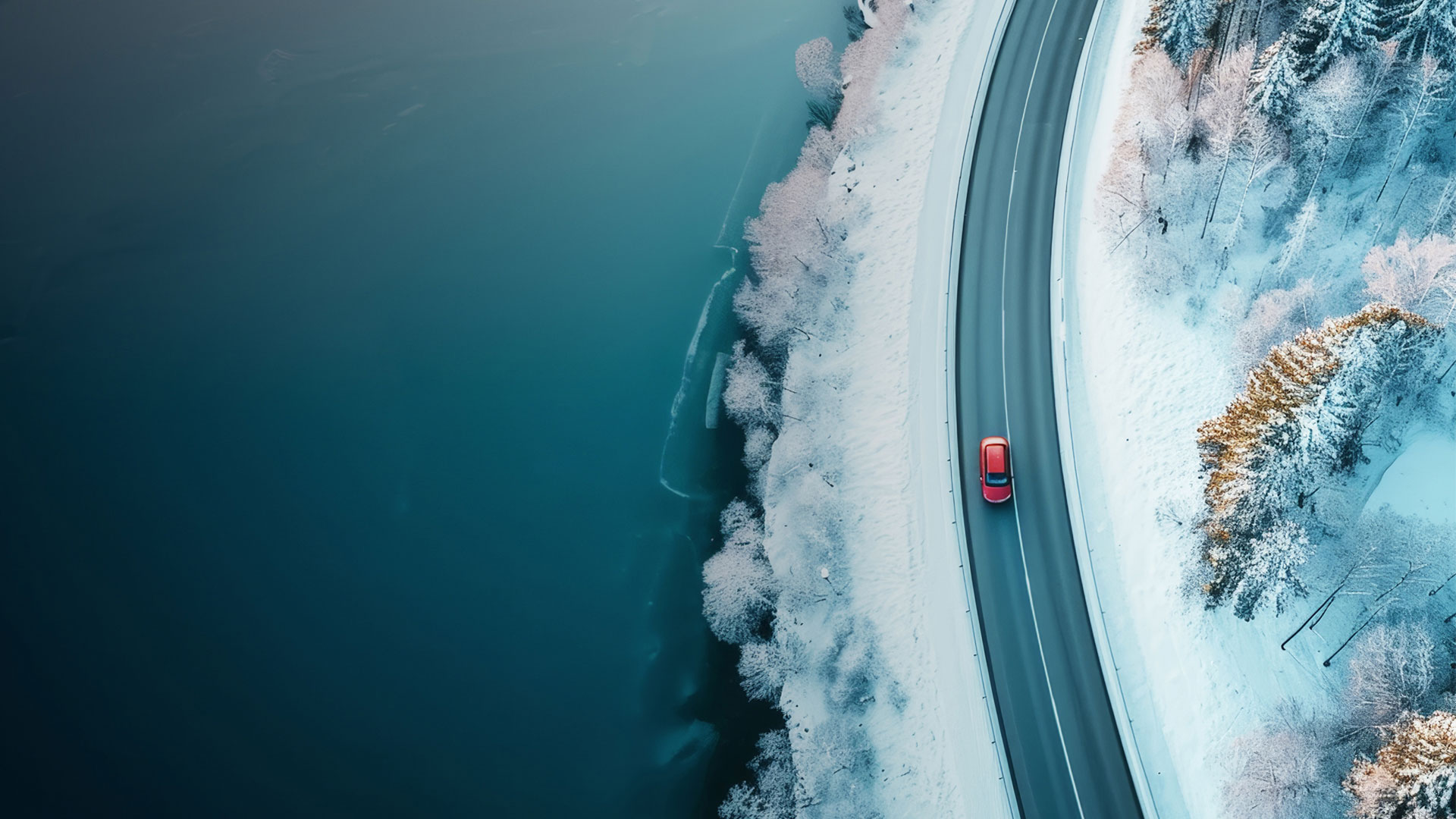 Dans un paysage enneigé, une voiture rouge circule sur une route en bordure d’un plan d’eau.